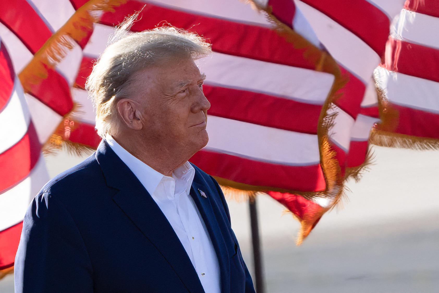 Trump is seen from the side, standing in front of a U.S. flag.