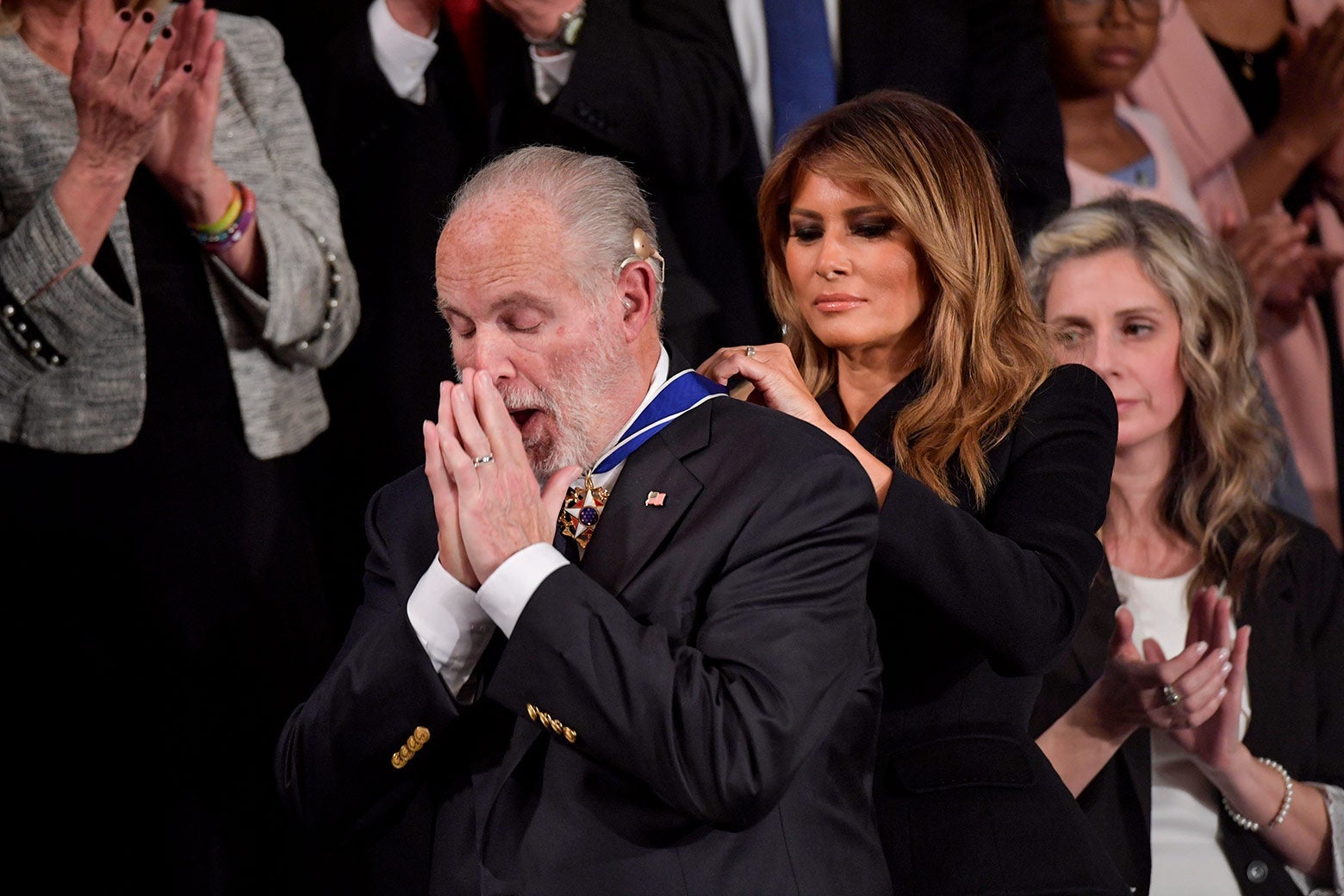 Melania Trump puts a medal around Rush Limbaugh's neck as he holds his hands to his face.