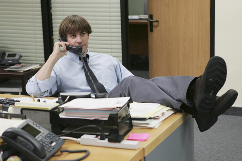 John Krasinki props his feet up on a desk as he talks on a landline phone.