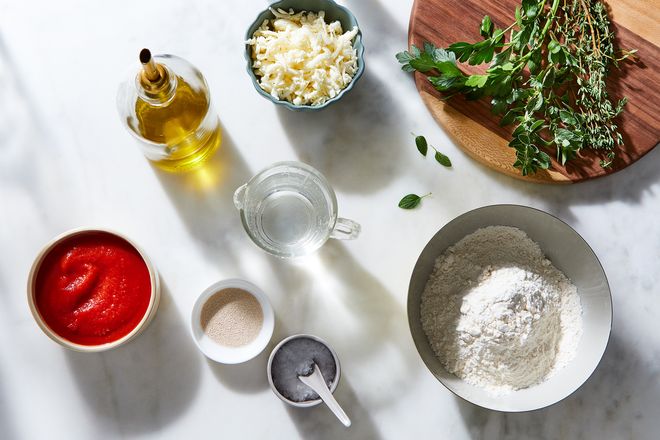 Small bowls of cheese, tomato sauce, olive oil, flour, and greens on a countertop.
