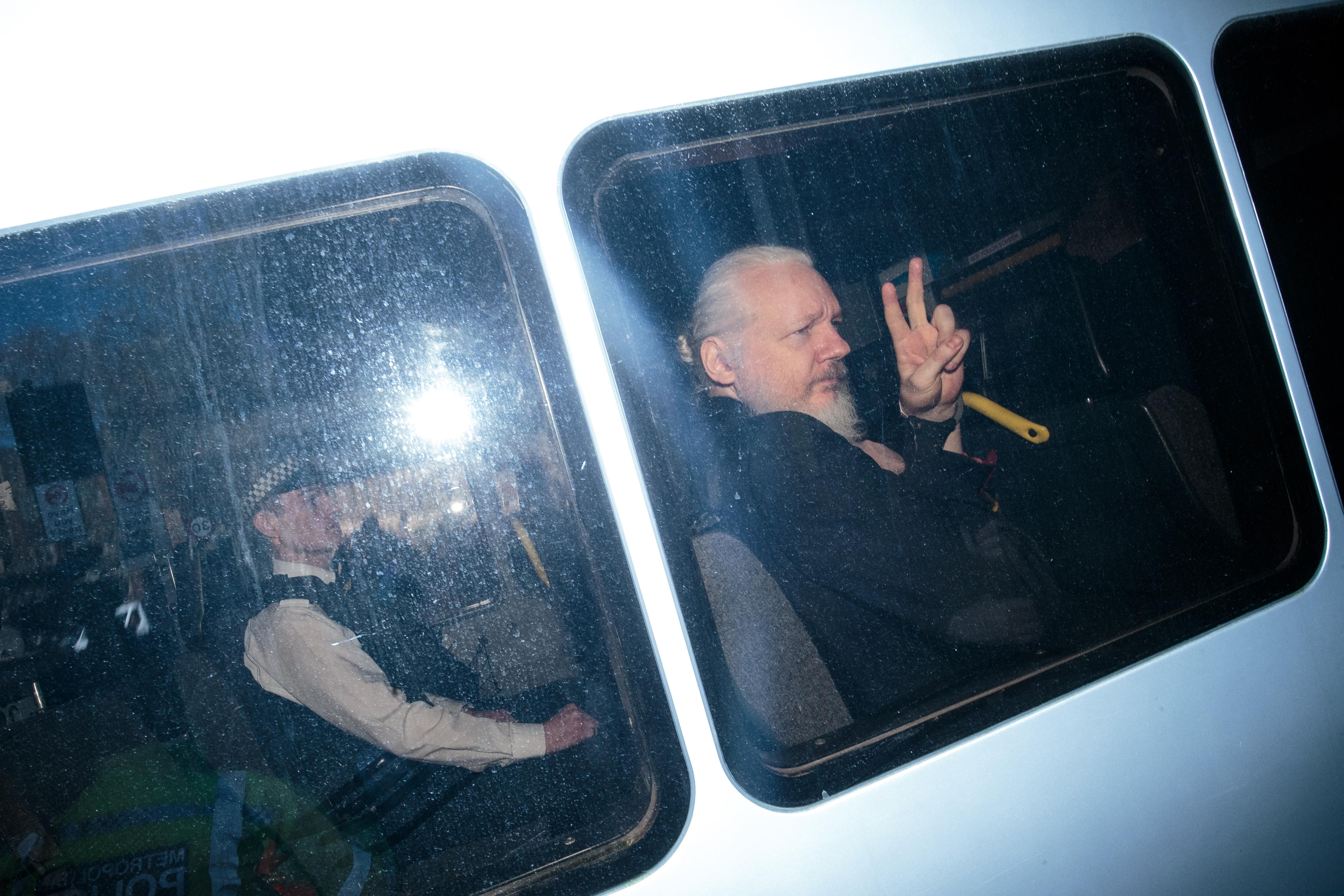 Julian Assange holds up a peace symbol as he is driven in a police vehicle in a grainy photo.