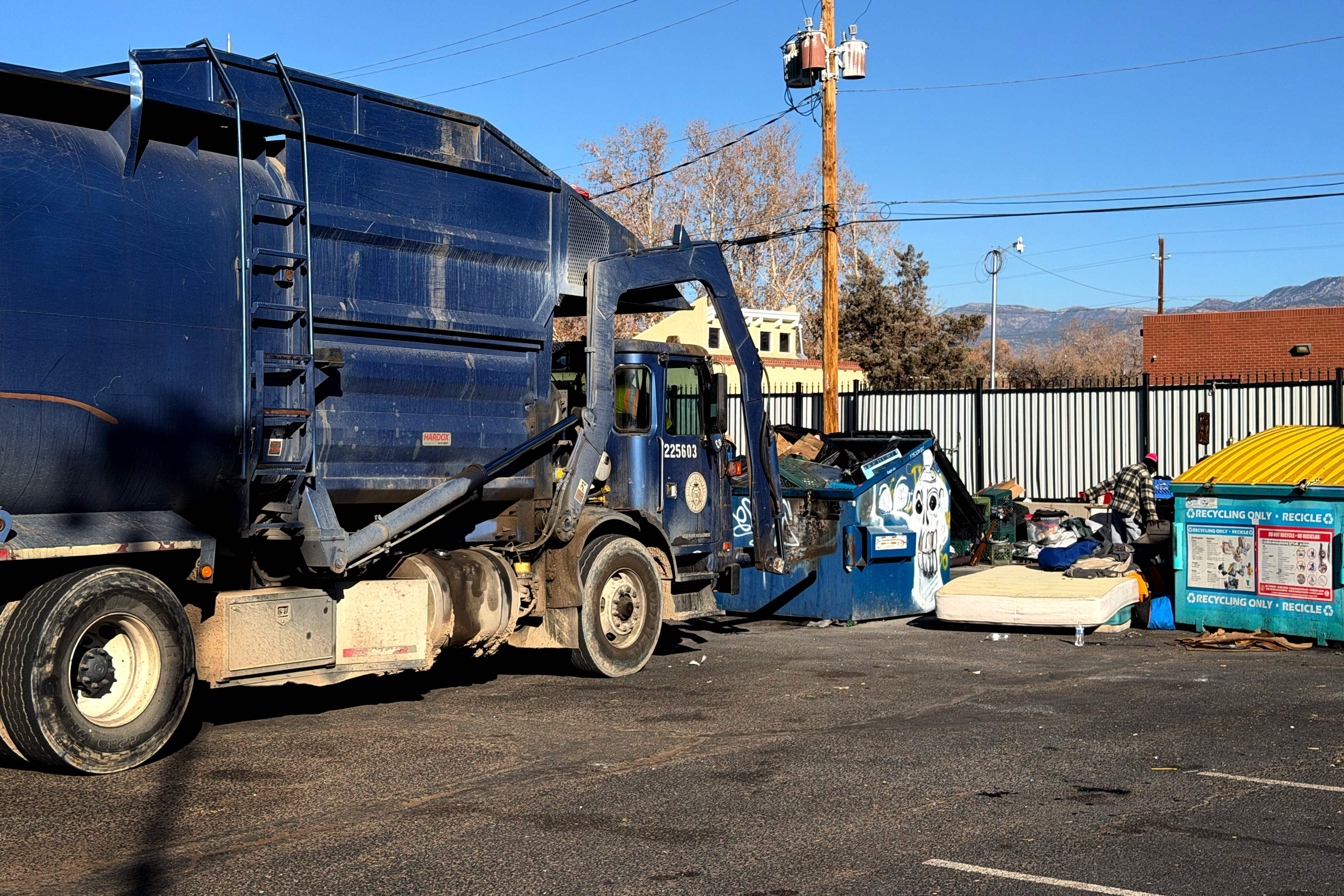 A garbage truck is picking up a full dumpster and there is a pile including a mattress next to it.