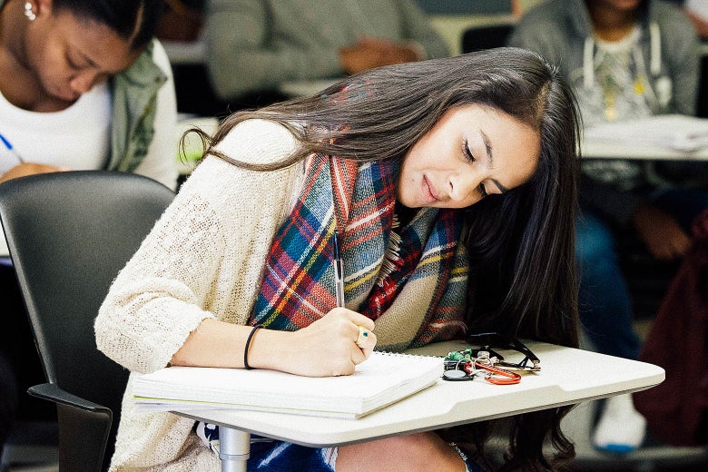 A young woman takes notes while seated at a desk in a classroom.