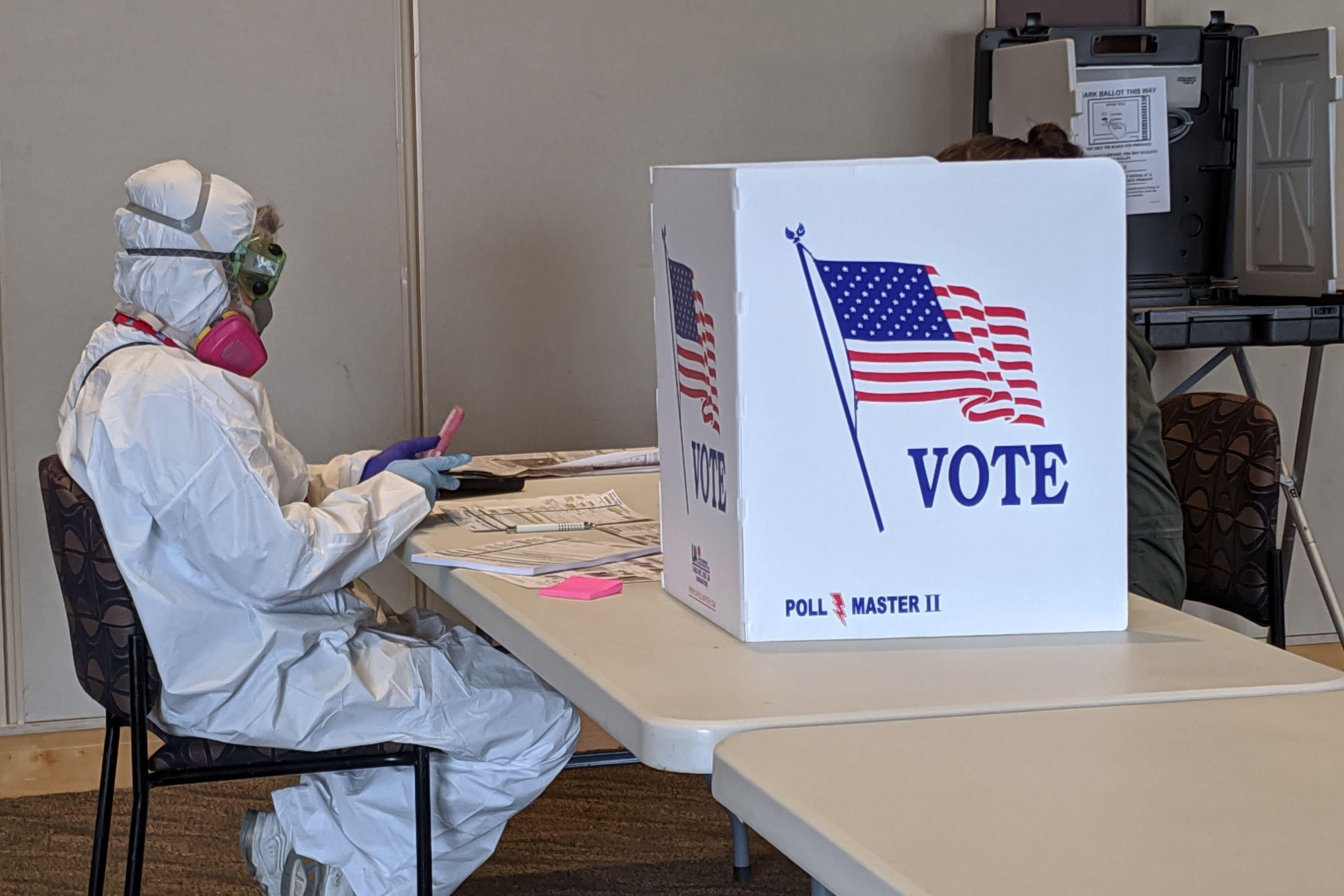 A person in a full-body hazmat suit sits at a table beside a voting booth