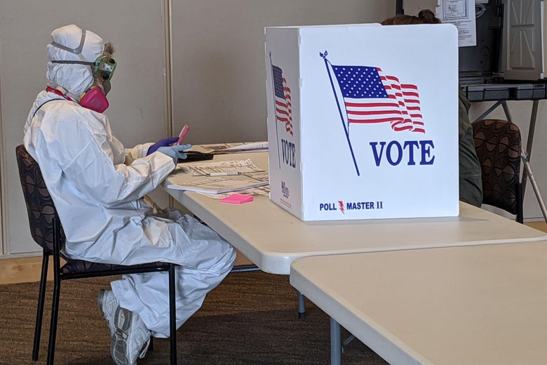A person in a full-body hazmat suit sits at a table beside a voting booth