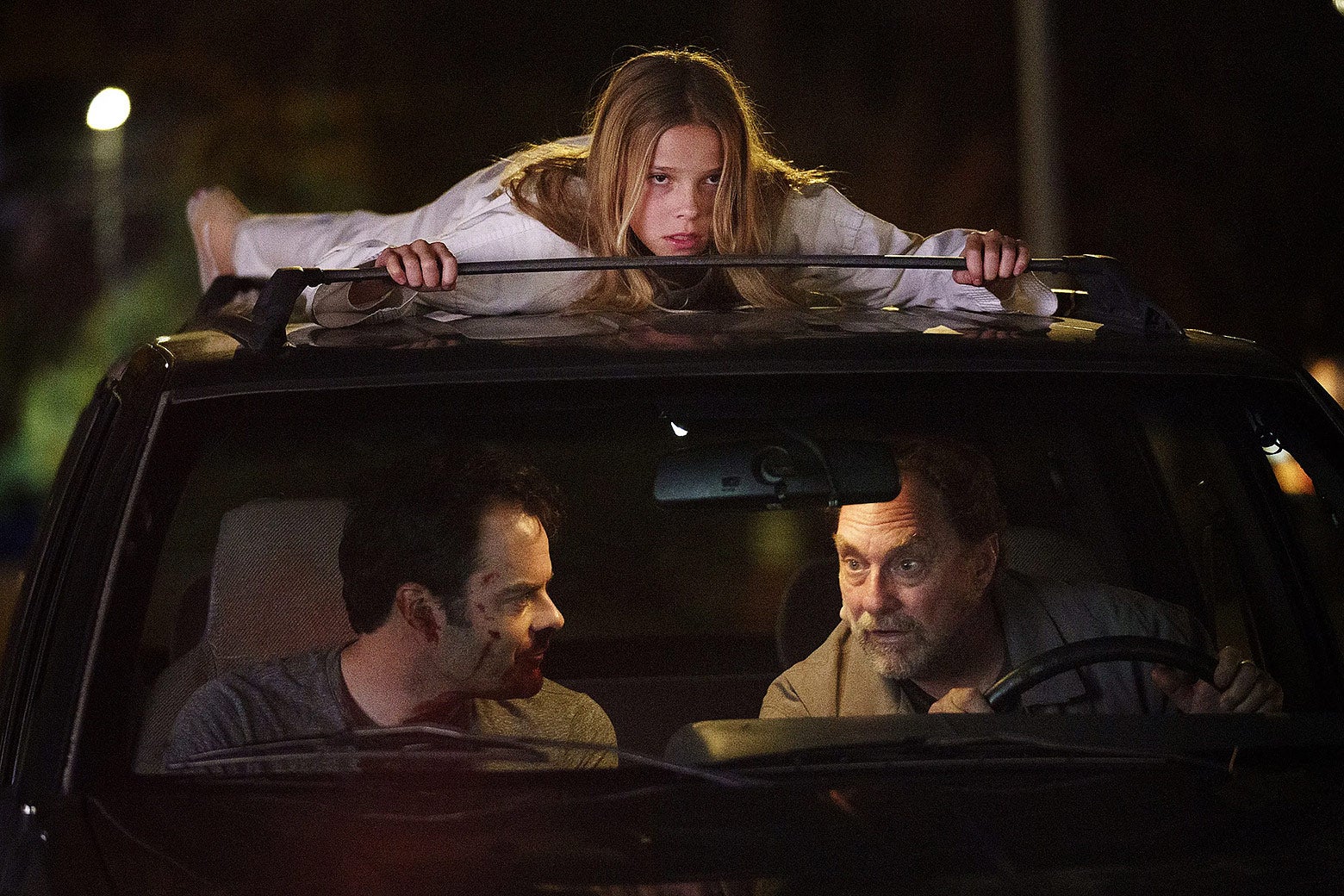 Barry and Ron sit inside a car, while a young girl in a white shirt perches atop it.