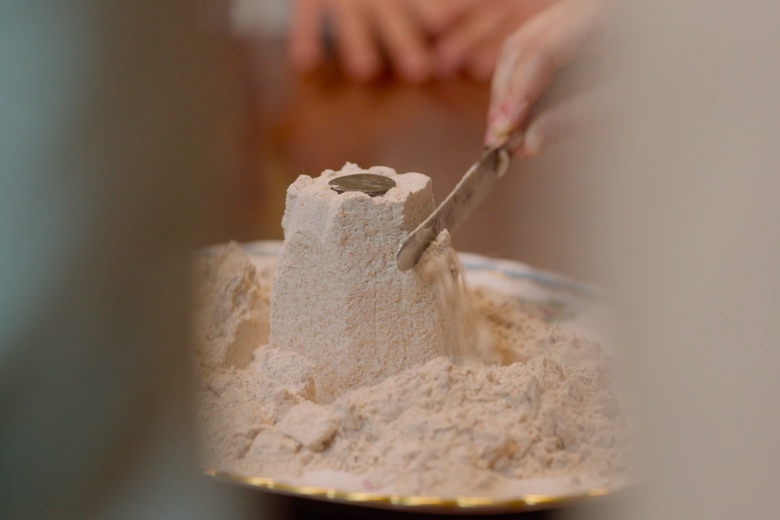 A narrow column of flour stands in a platter full of flour, topped with a coin and with a butter knife about to cut into the flour.