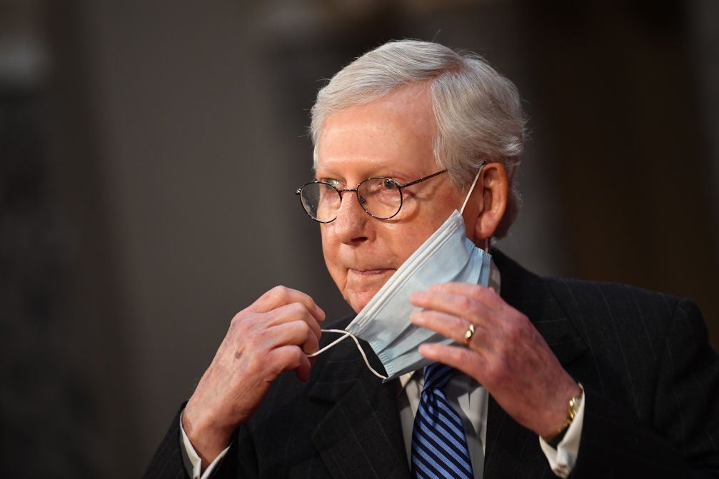 Mitch McConnell, seen from the shoulders up, gazes to his right as he removes a blue surgical mask from his right ear, with it still dangling from his left ear.