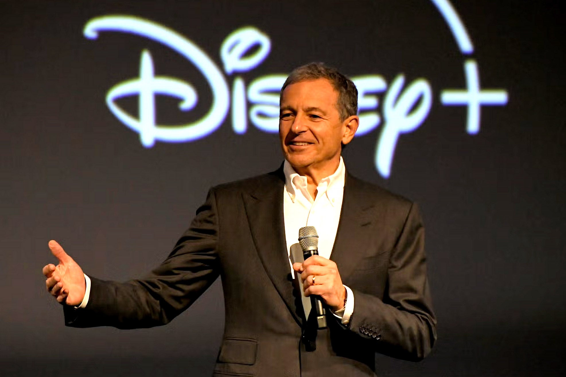 A middle-aged white man in a suit smiles and holds a microphone onstage in front of a backdrop that says "Disney."