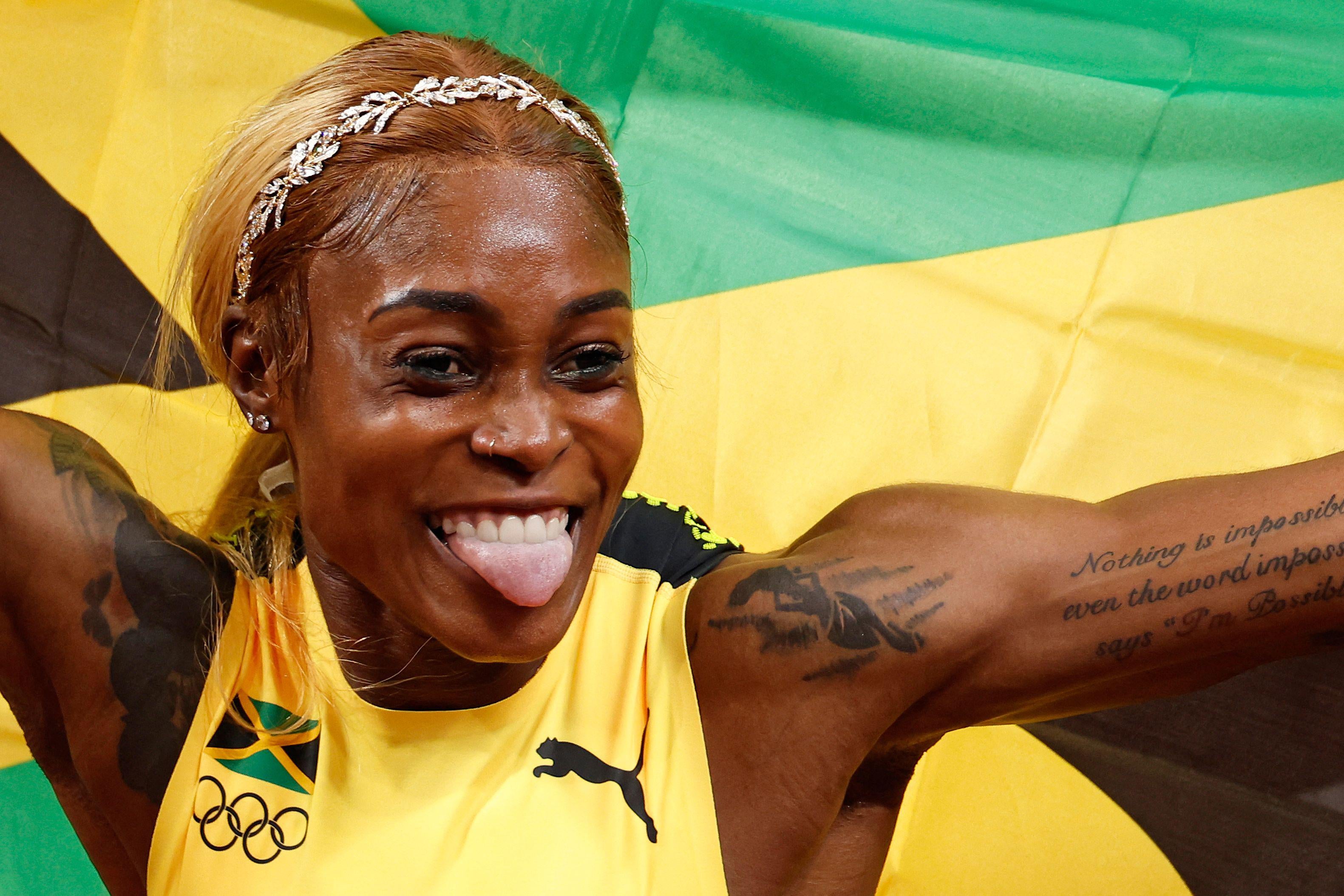 Thompson-Herah sticks her tongue out and smiles, holding a large Jamaican flag behind her, in her jersey and revealing tattoos on both arms