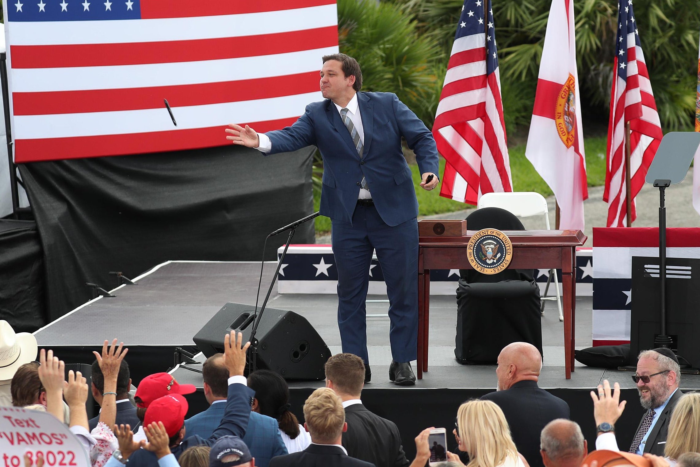 Ron DeSantis stands at the front of a stage with a desk behind him. He throws a black Sharpie into the crowd of people below, their hands outstretched.