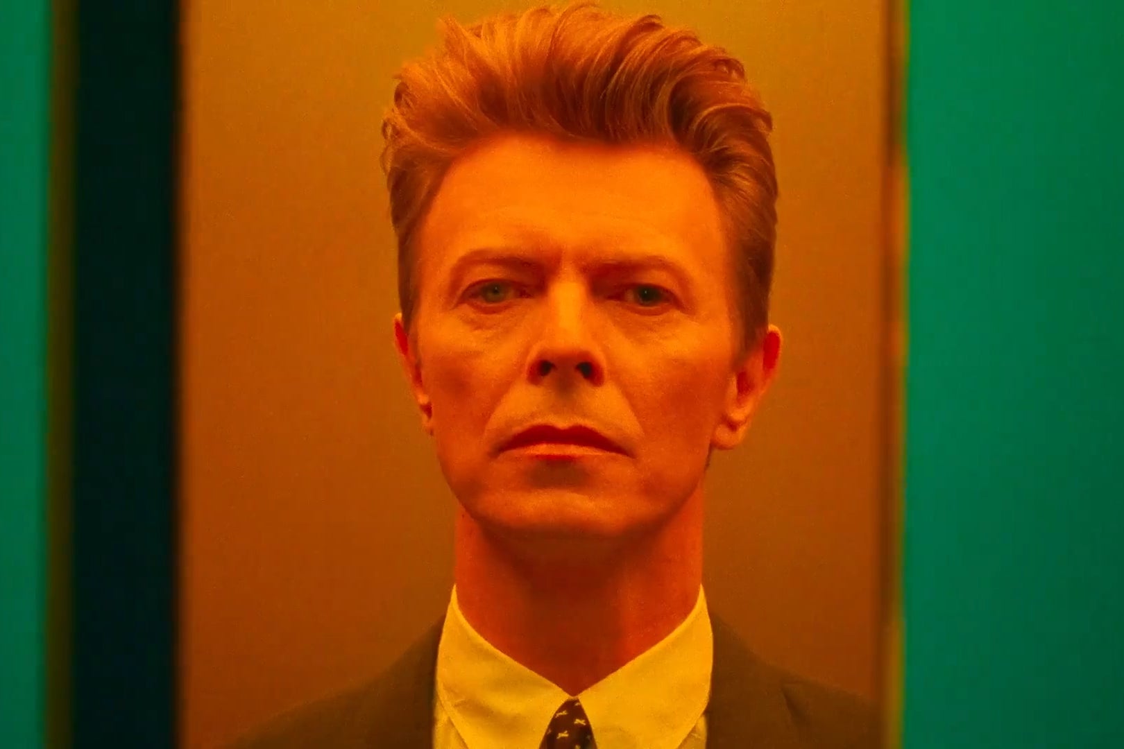 A close up of David Bowie, awash in orange-tinged light, as he stands in a suit jacket and tie and stares deadpan out into the distance. 