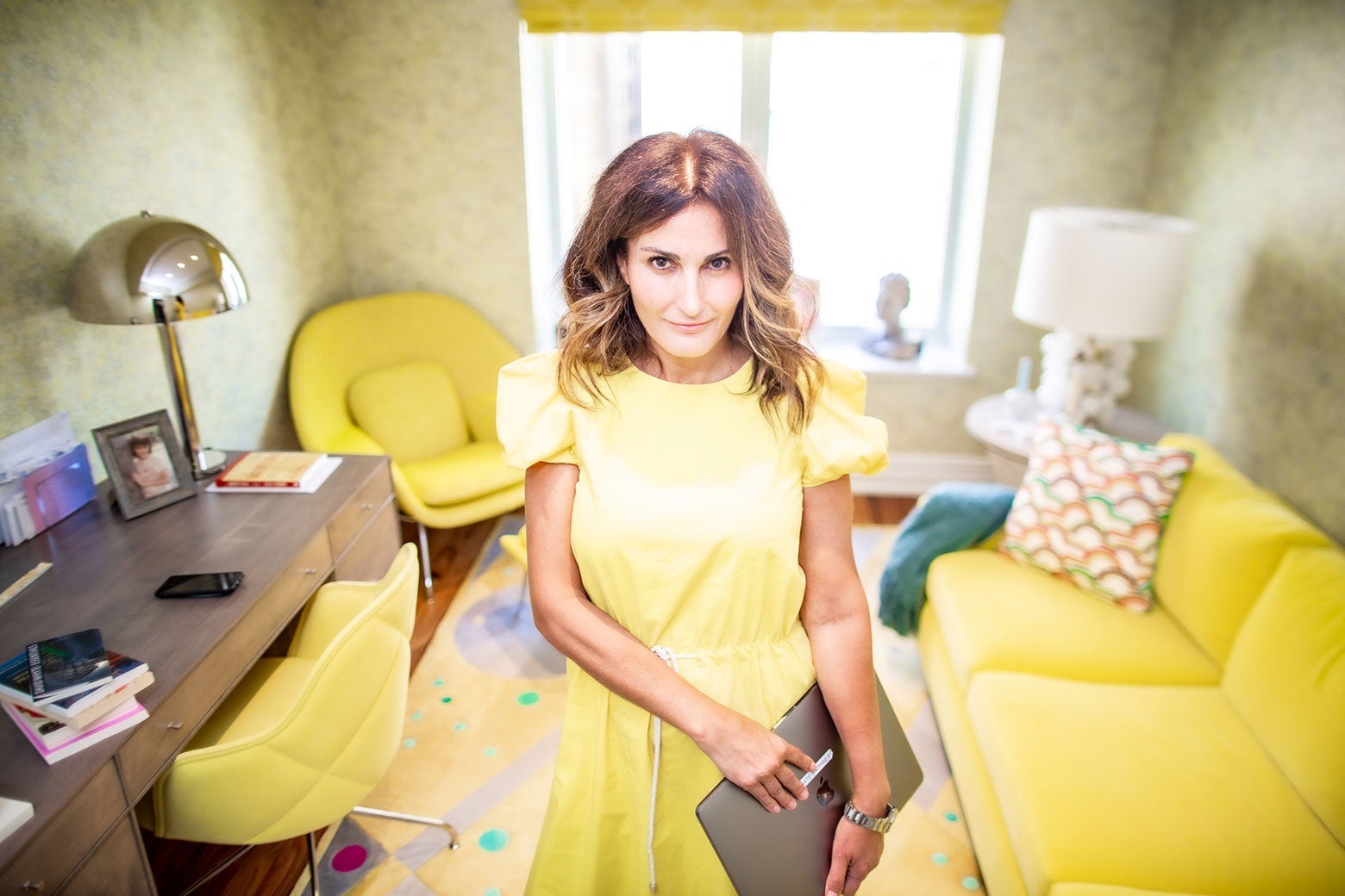 A woman in a yellow dress holds an Apple laptop while standing in a yellow room. 