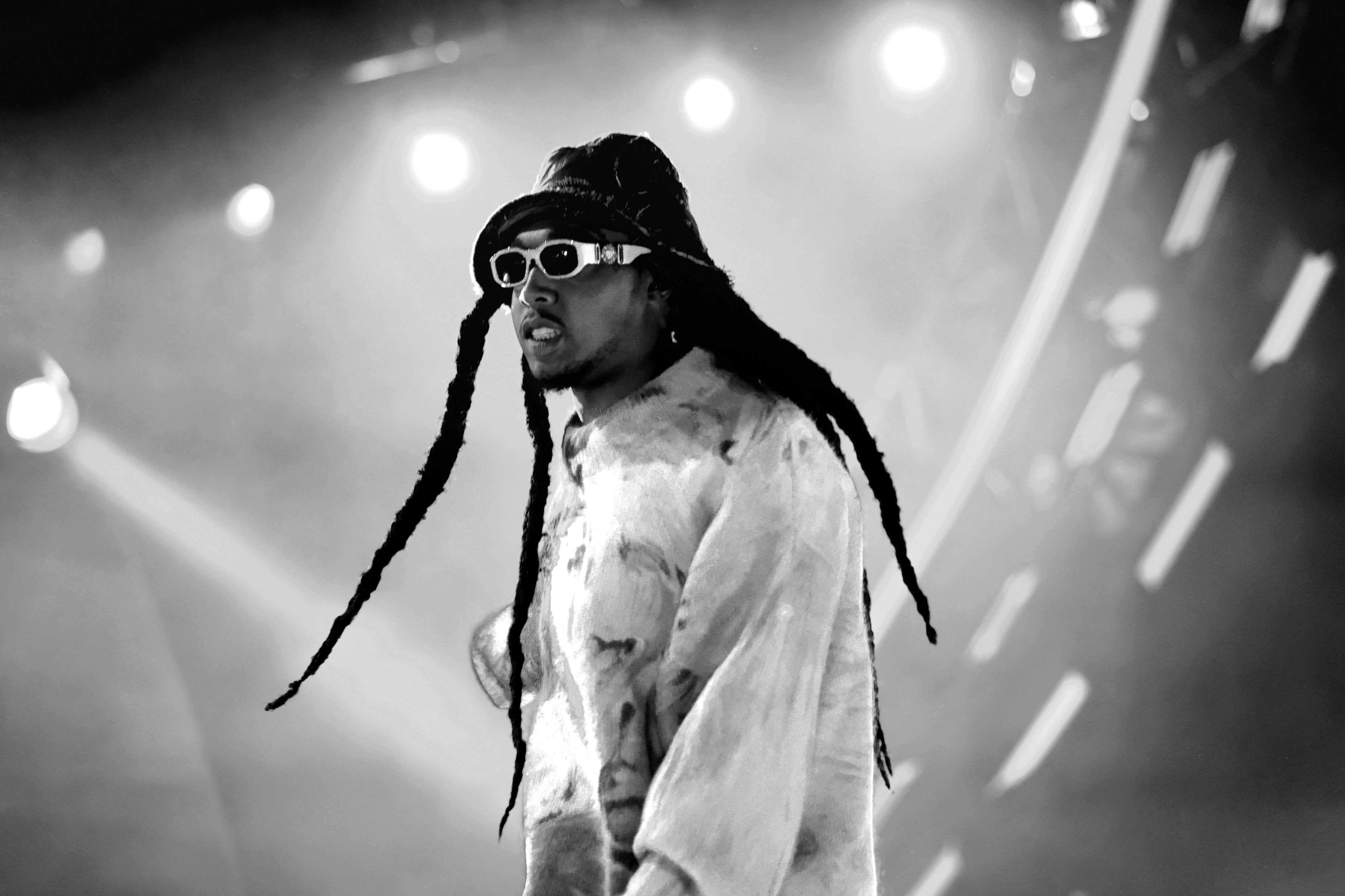 A black-and-white photo shows the rapper looking stylish on stage in sunglasses and an almost Paddington-esque hat and a baggy patterned shirt or sweatshirt, his teeth bared and his long dreads flying out around him.