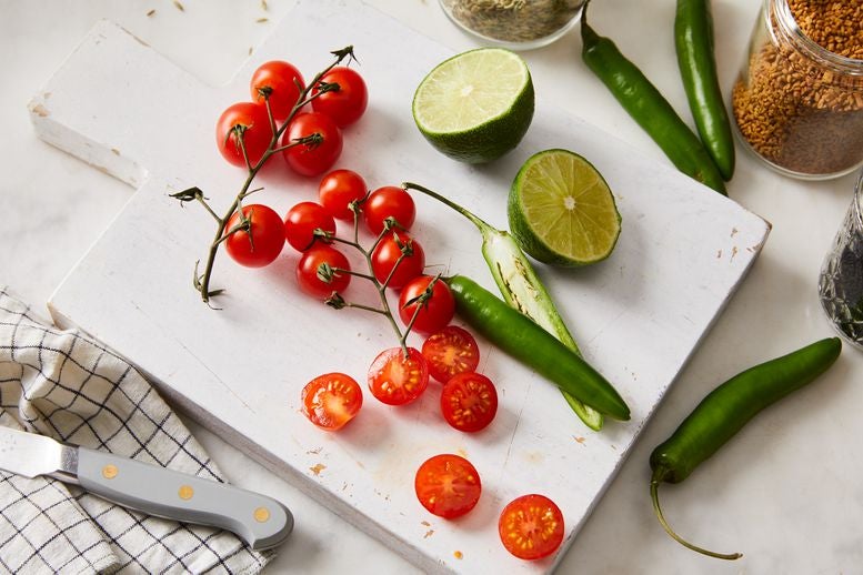 Cherry tomatoes, a halved lime, and green peppers on a cutting board with jars of spices next to them.