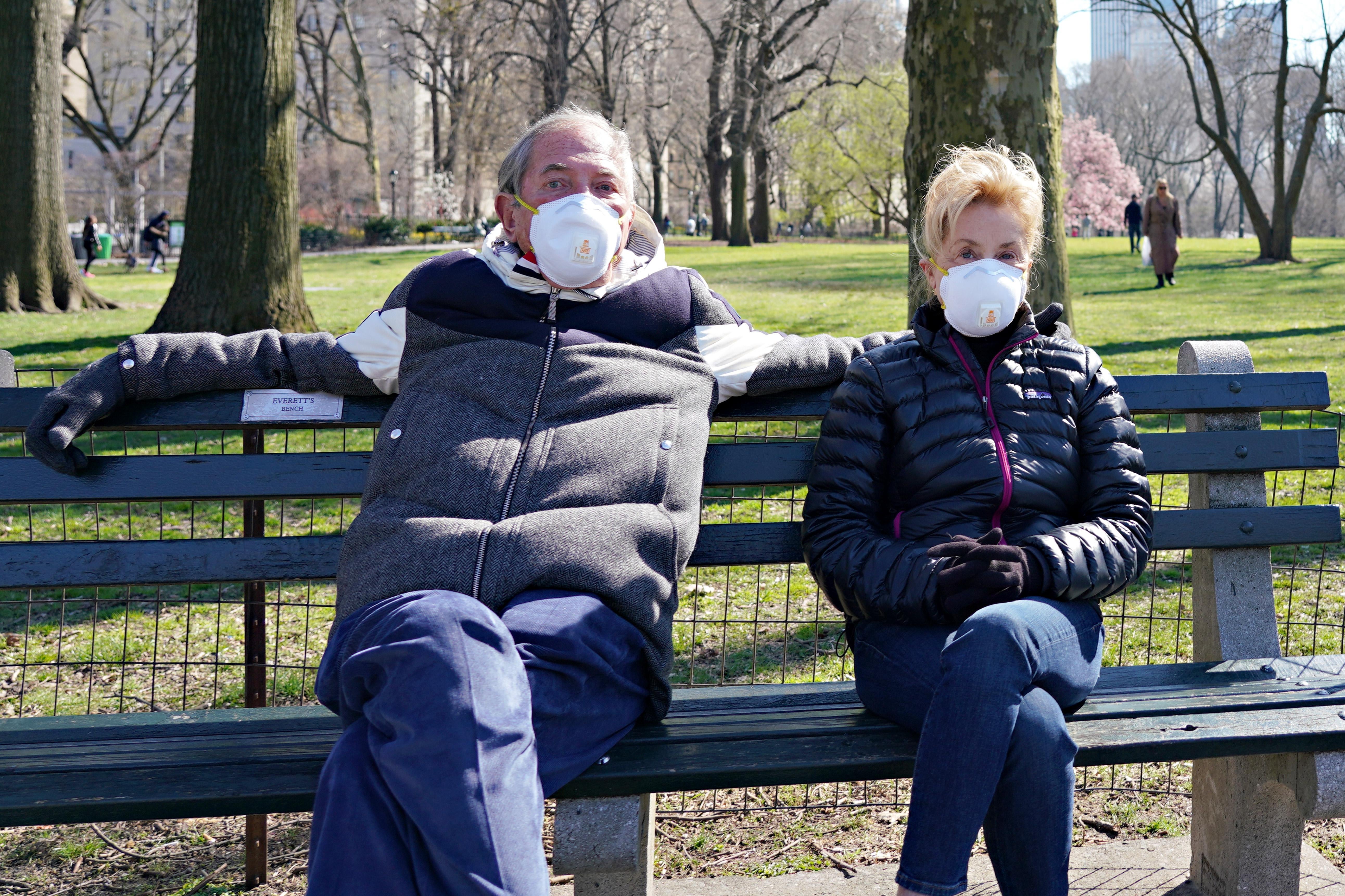 A man and a woman, both wearing masks, sit on a bench in the park