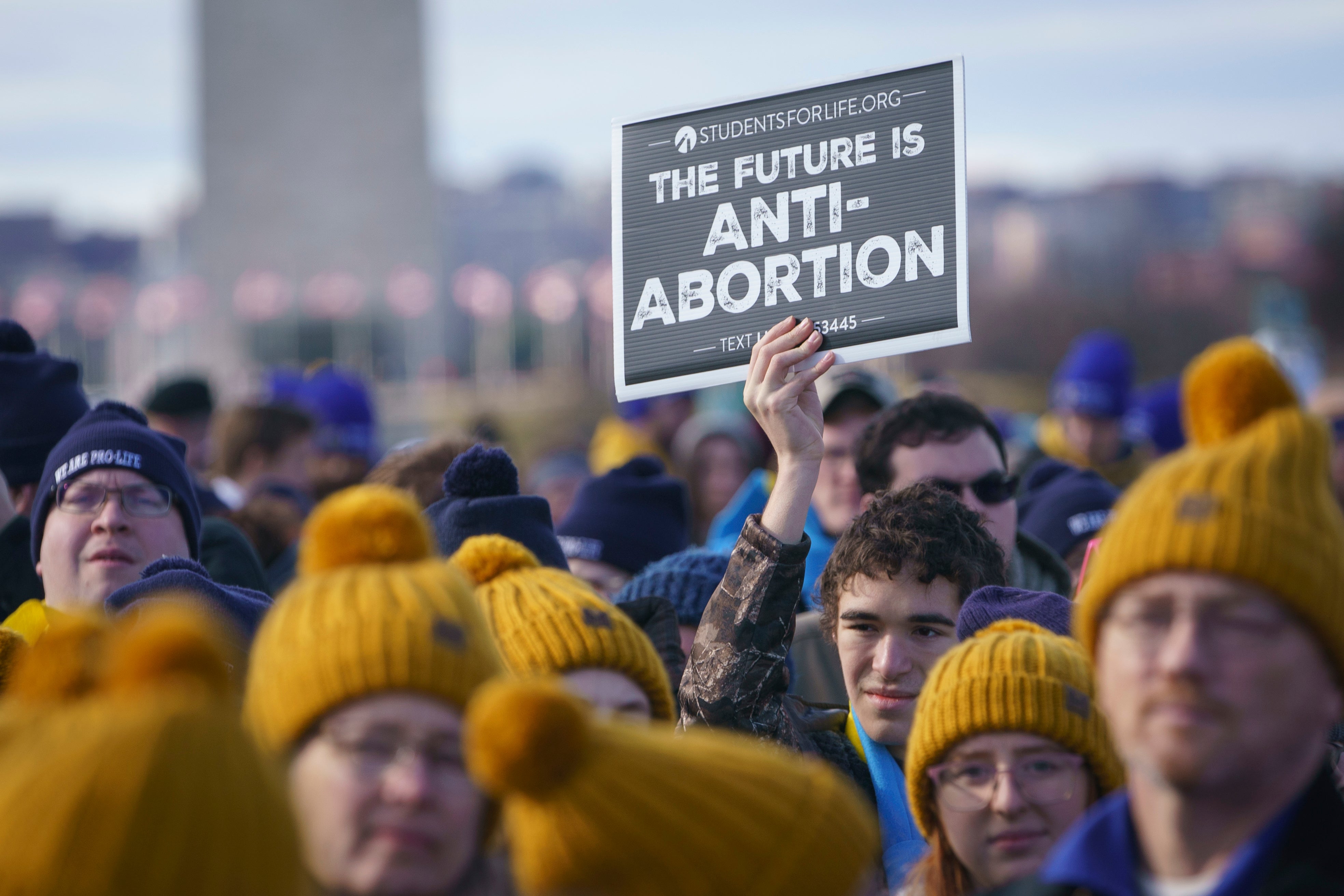 A photo of people wearing yellow hats, with a young man behind them holding a sign that reads: "The Future Is Anti-Abortion."