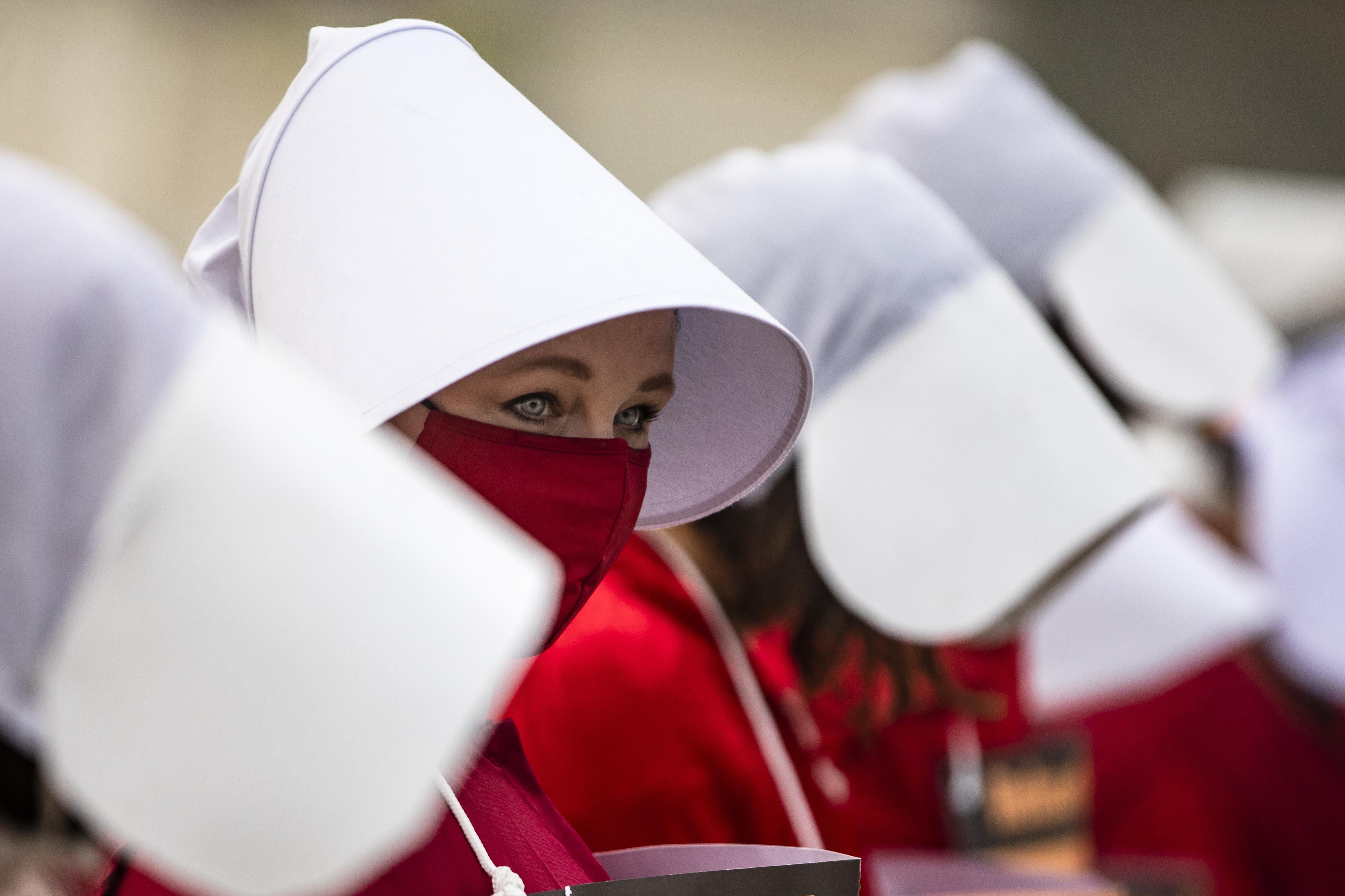 In a line of women wearing large white bonnets, one woman peeks over her red mask.