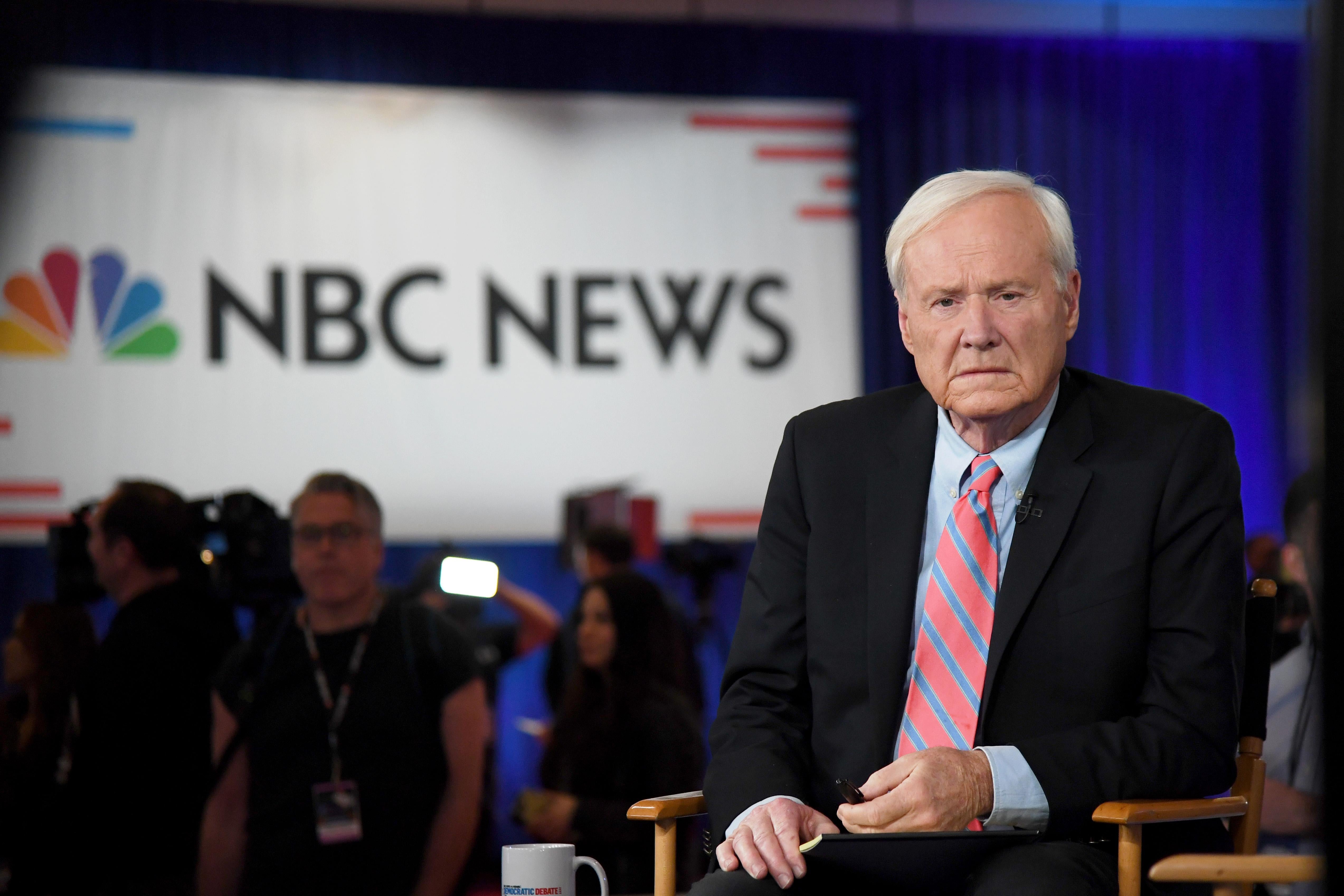 Chris Matthews seated, looking serious, before going on the air, with the NBC News logo on a screen in the background.