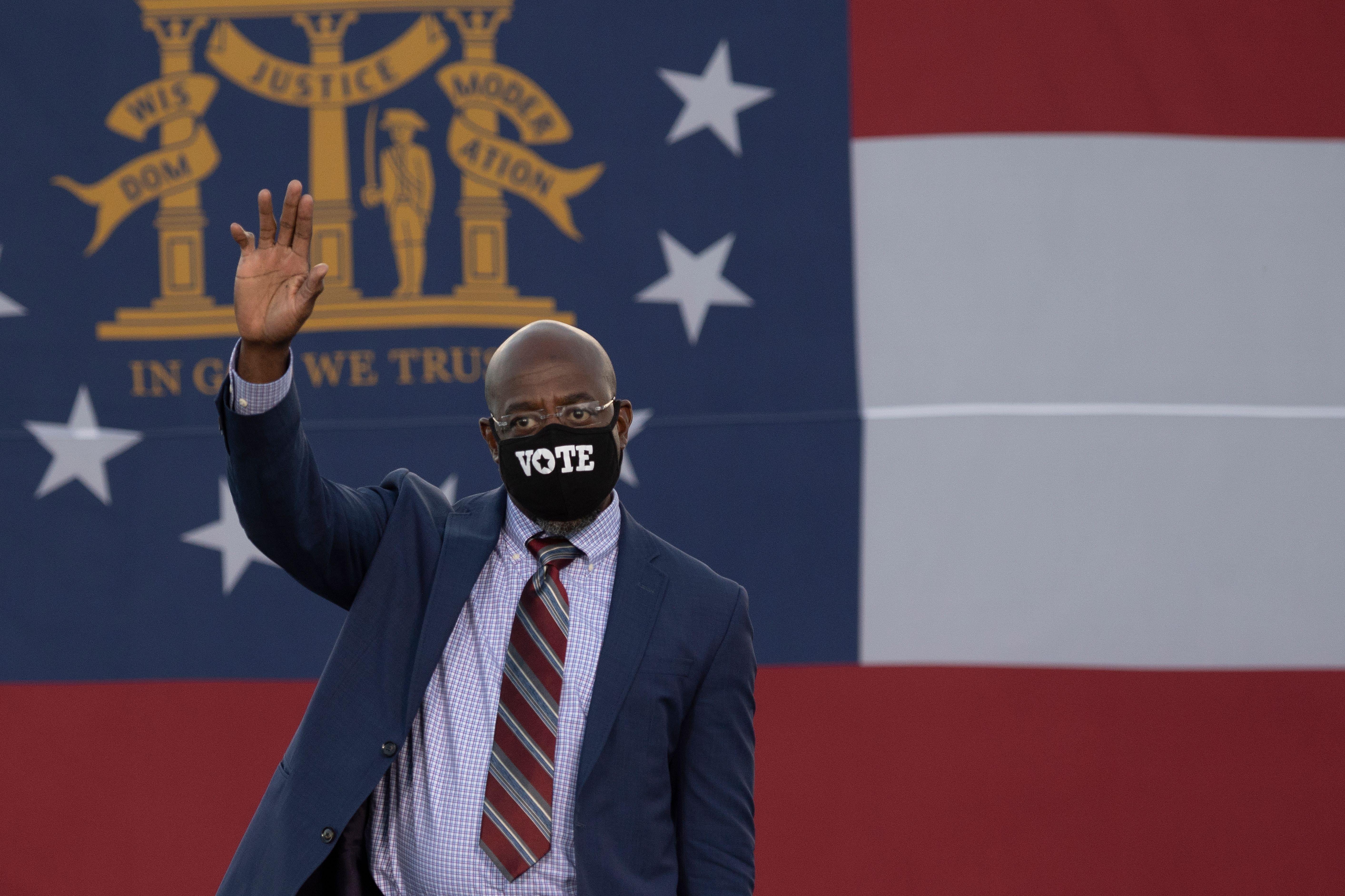 Raphael Warnock waves during a rally while wearing a mask that reads "vote" and standing in front of the Georgia state flag. 
