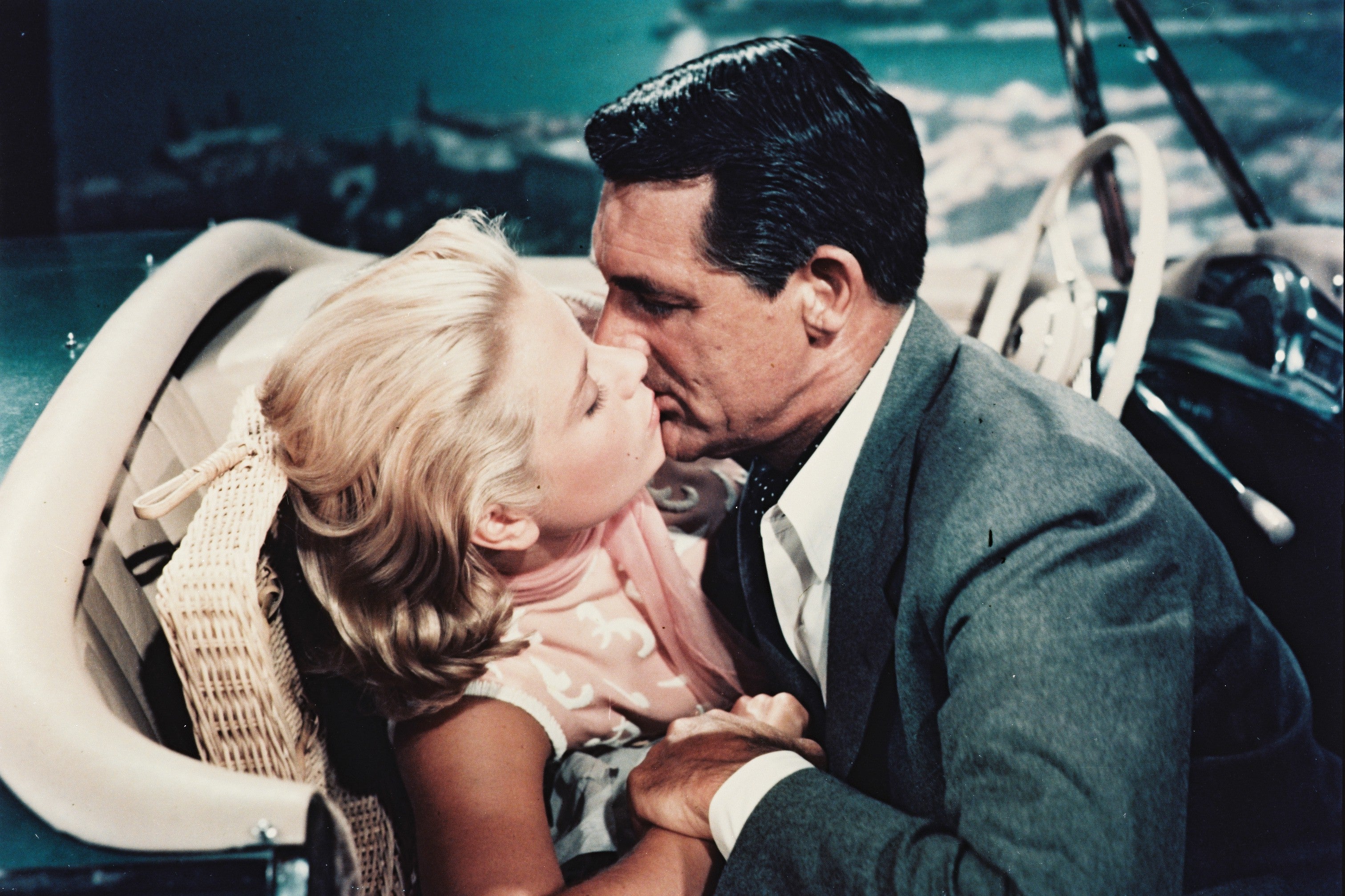 In this still from the classic 1950s movie: Cary Grant in a gray suit gives Grace Kelly, donning a pink dress, a passionate kiss while they're in a classic car. 