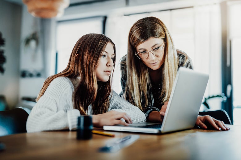 A girl and her mother sit a table and look at a laptop screen.