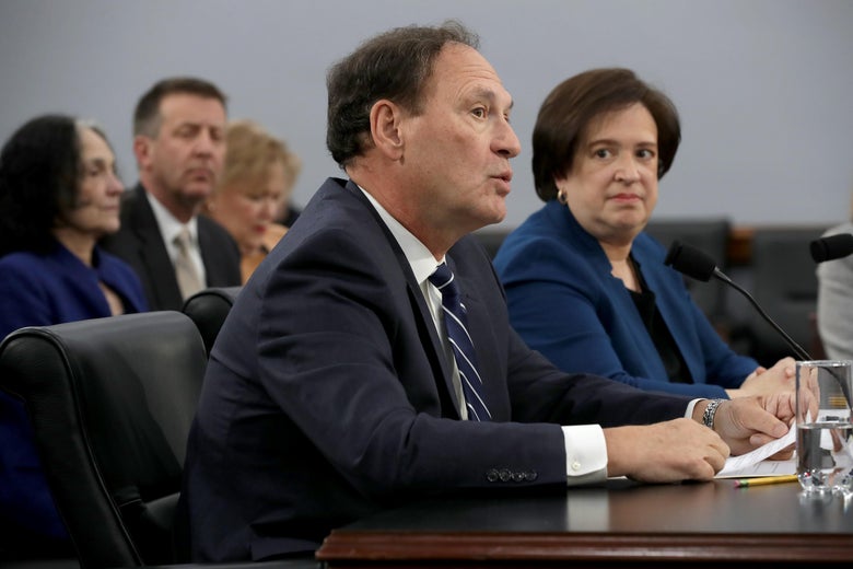 Alito speaks into a mic while seated at a table in a hearing room. Kagan looks on, sitting beside him.