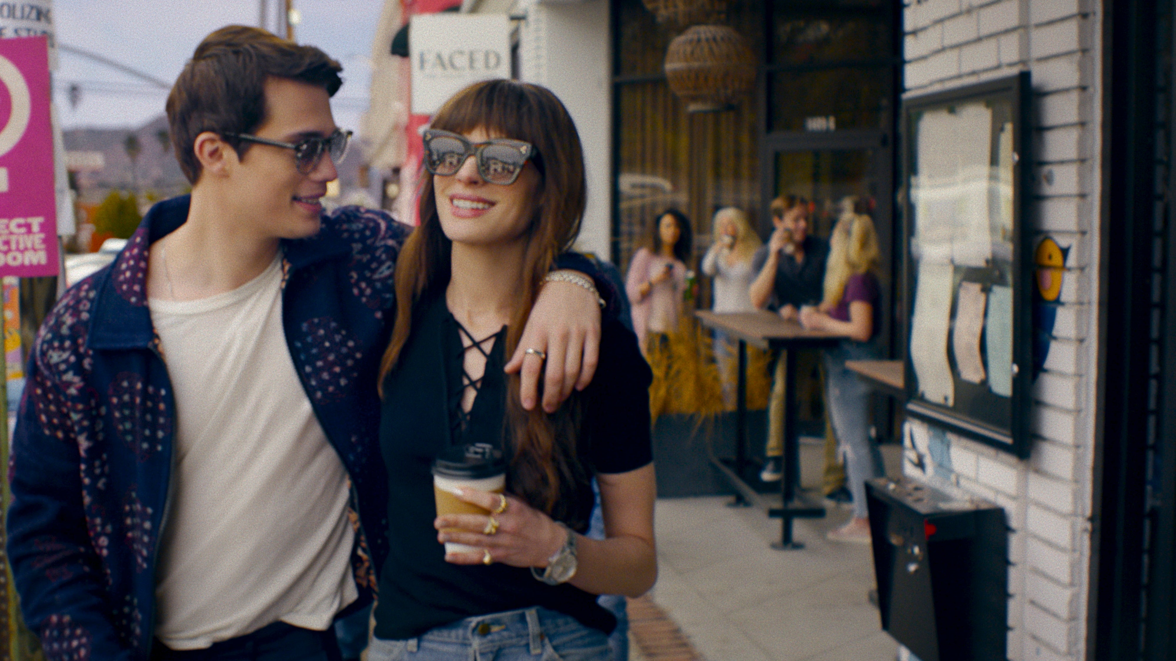 Galitzine and Hathaway walk down a busy street holding coffees and wearing sunglasses; his arm is over her shoulders.