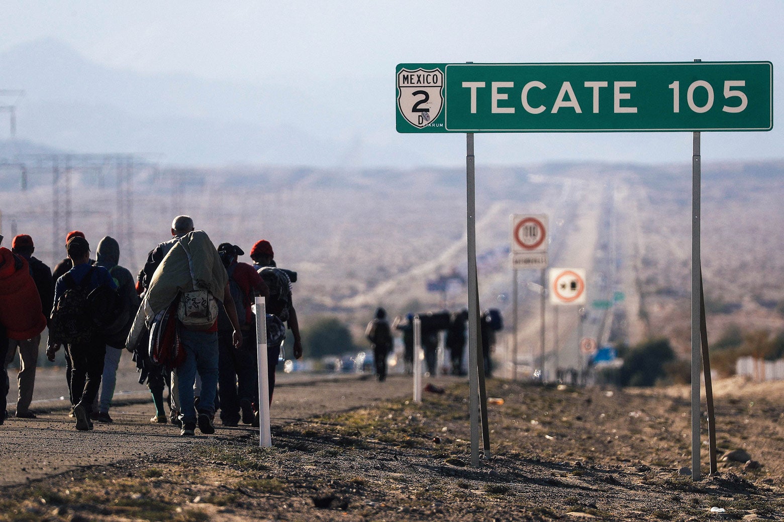 Migrants walk down a road beside a road sign that says Tecate 105.