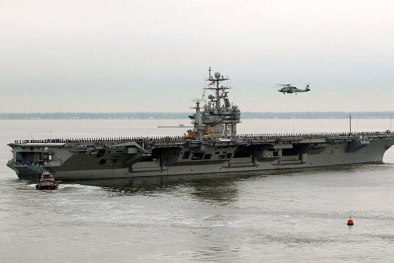 An aircraft carrier on the water, trailed by a helicopter above.