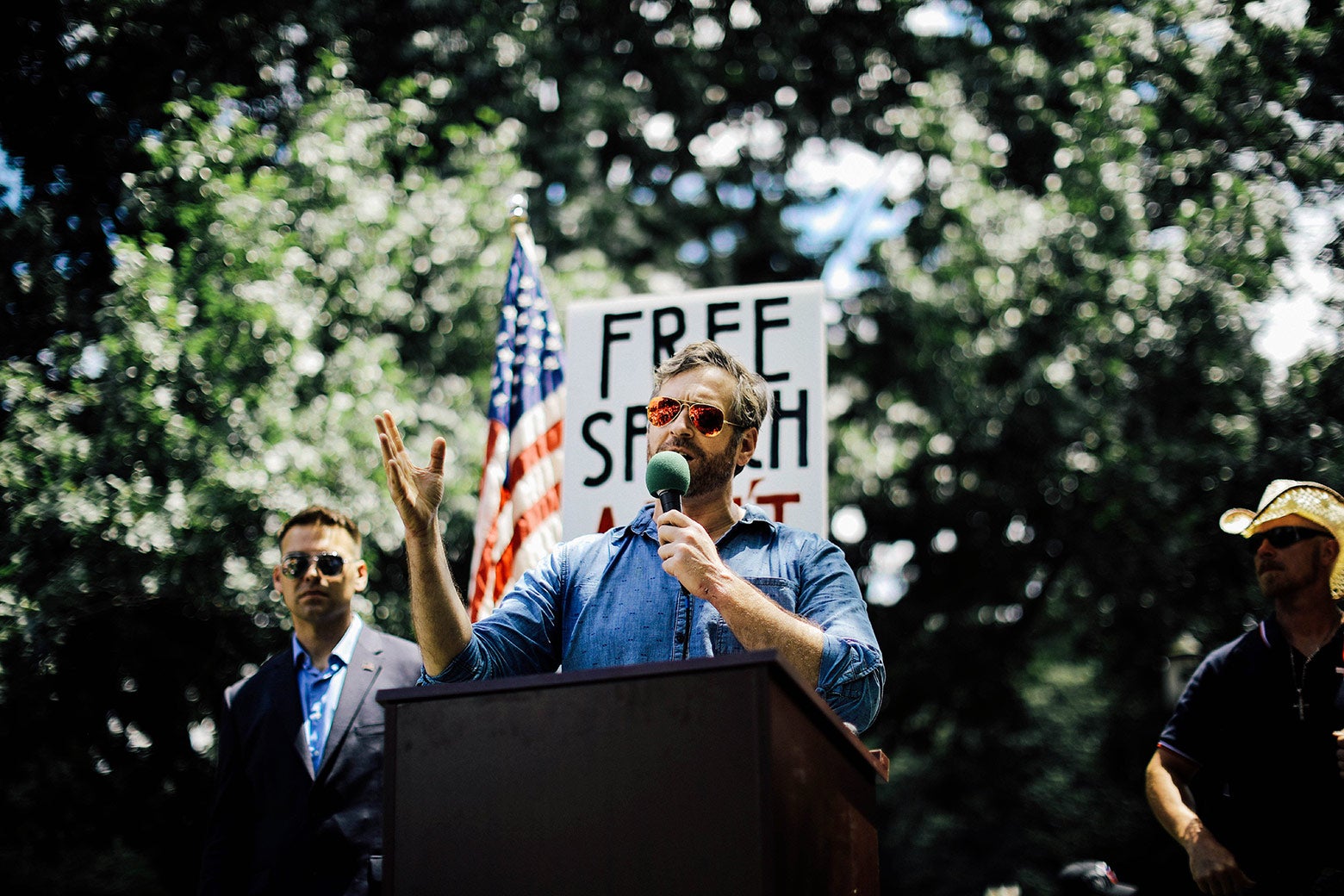 Mike Cernovich speaks during a rally with a sign that says "Free Speech" in the background.