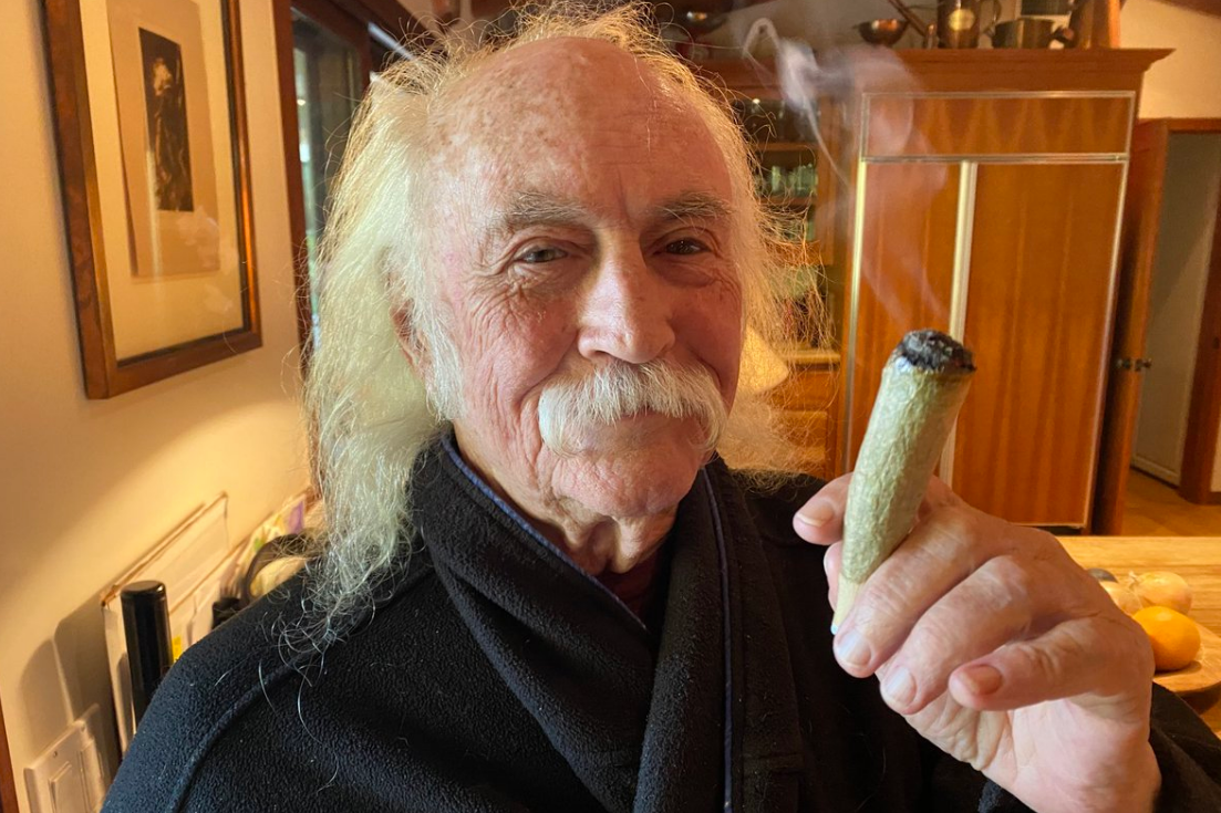 An aging David Crosby in a white handlebar mustache and a black robe smiles while holding up a truly humongous joint
