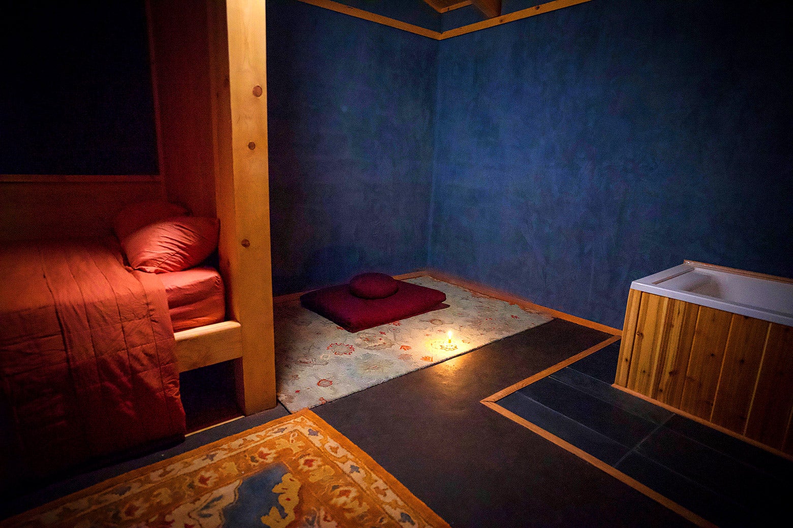 A bed, rug, bathtub, and meditation pillow in a blue-painted room are lit by what appears to be a single candle on the floor by the pillow.