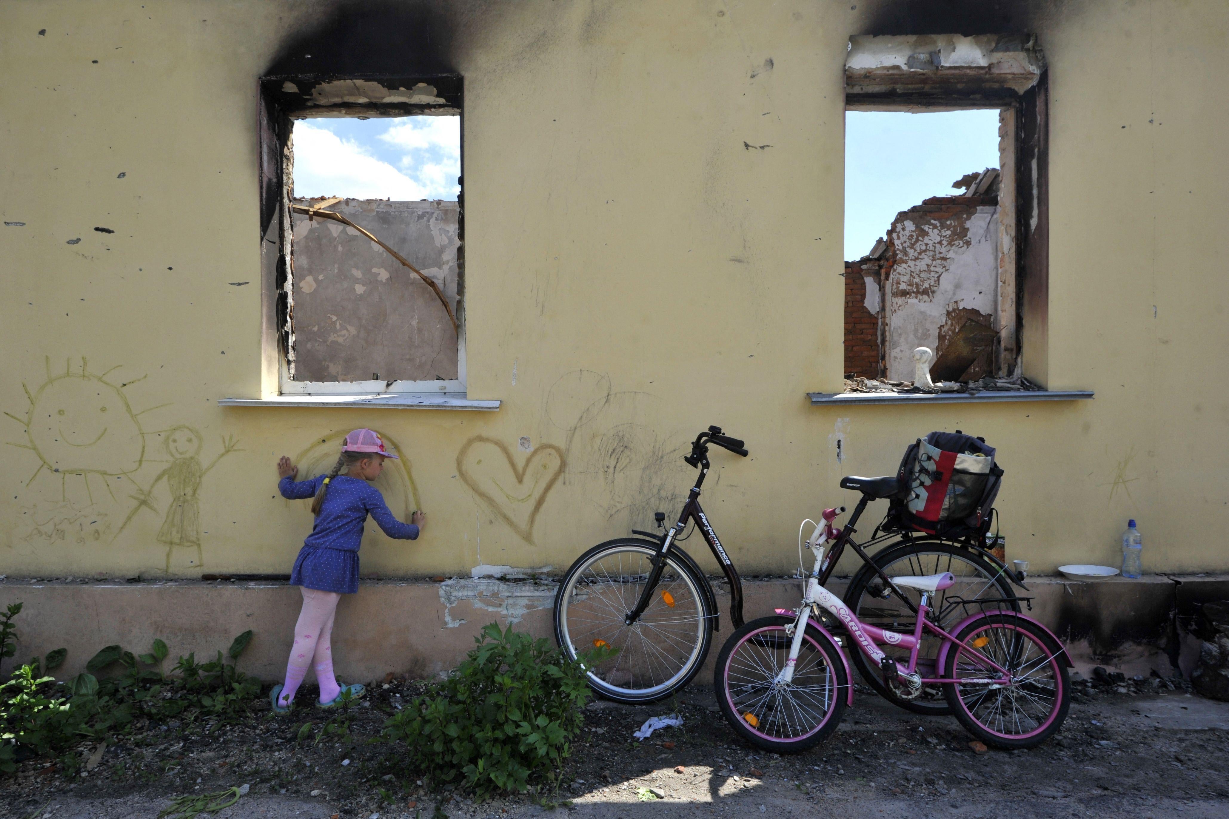 A young girl draws on the wall of a destroyed house with bicycles nearby