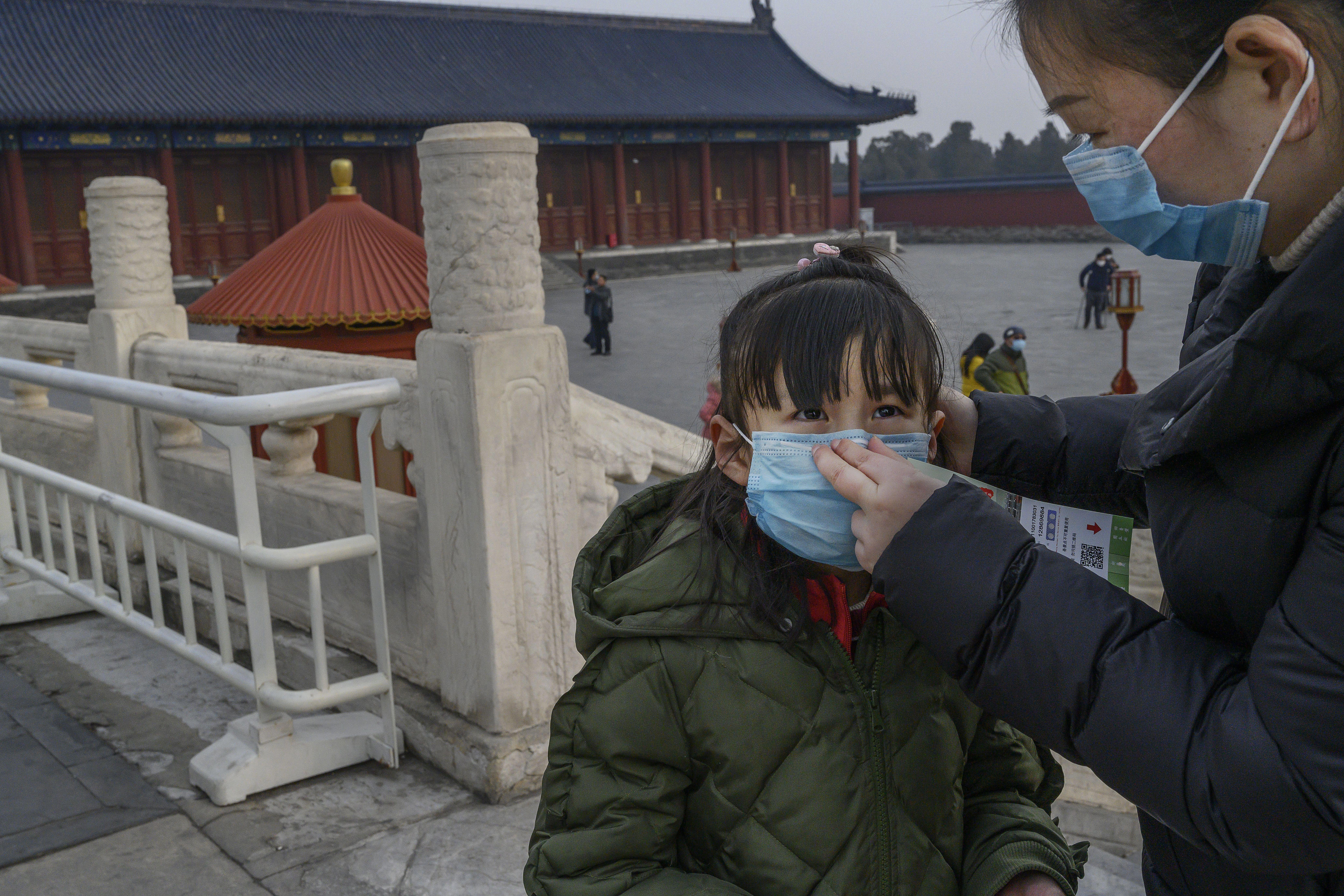A woman with a protective mask places a mask on a young girl to her left in front of a red temple.