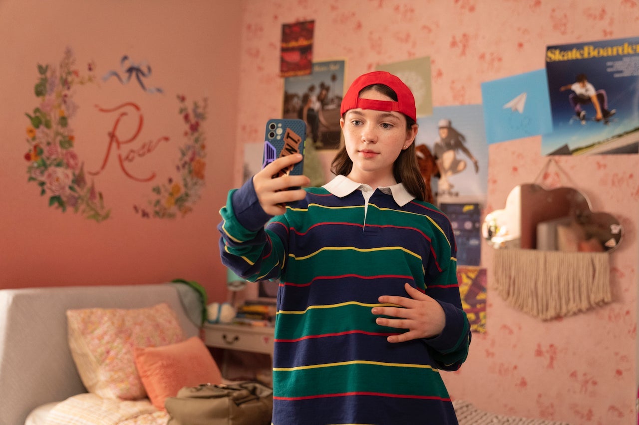 A teen stands in their room wearing a red backward cap and green-and-blue striped shirt. They look at their phone with their hand outstretched, taking a selfie in their bedroom.