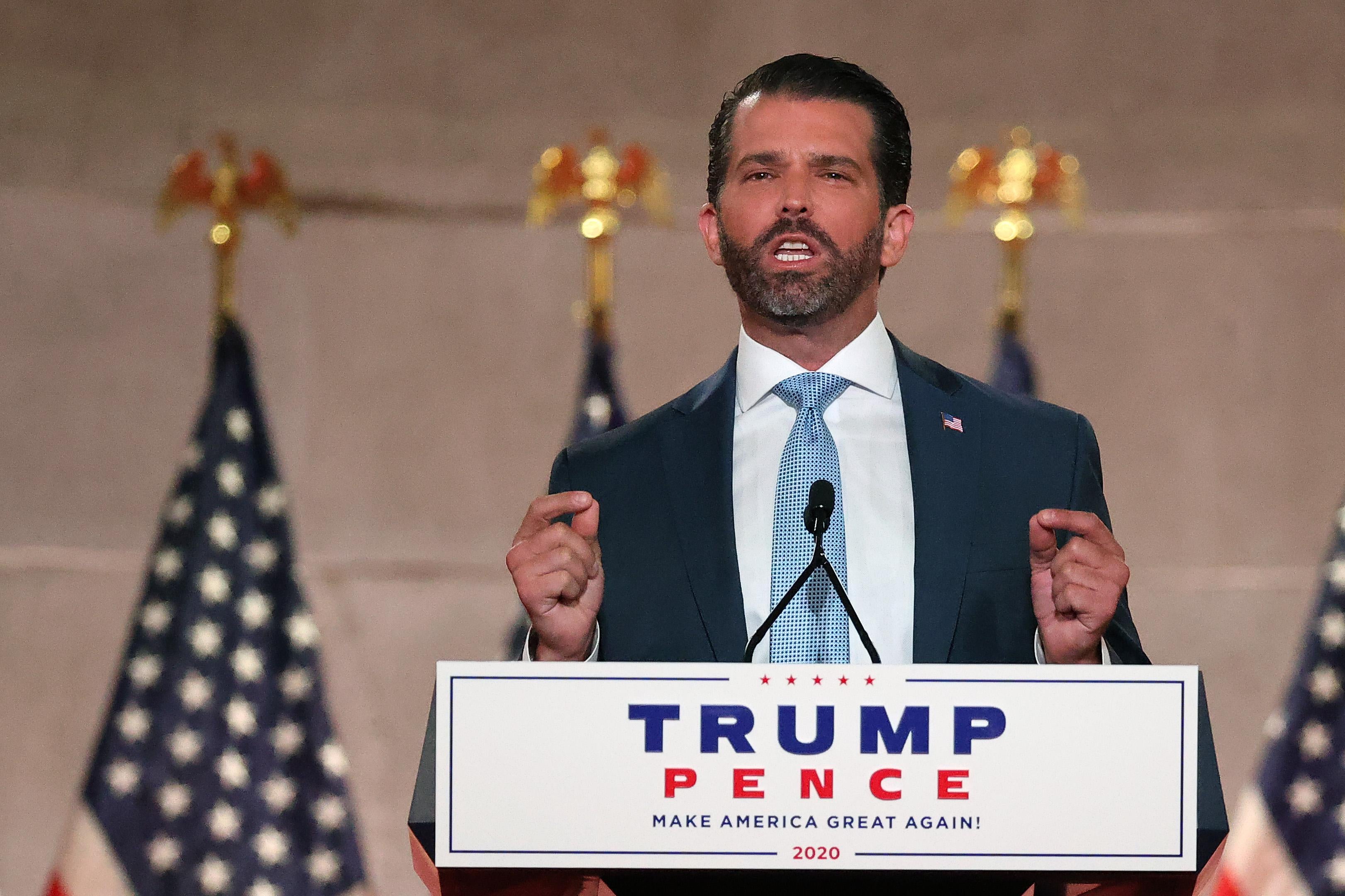 Donald Trump Jr. stands behind a podium with a "Trump Pence" sign on it. He gestures with both hands, touching his pointer fingers and thumbs together. Several U.S. flags stand on poles behind him.