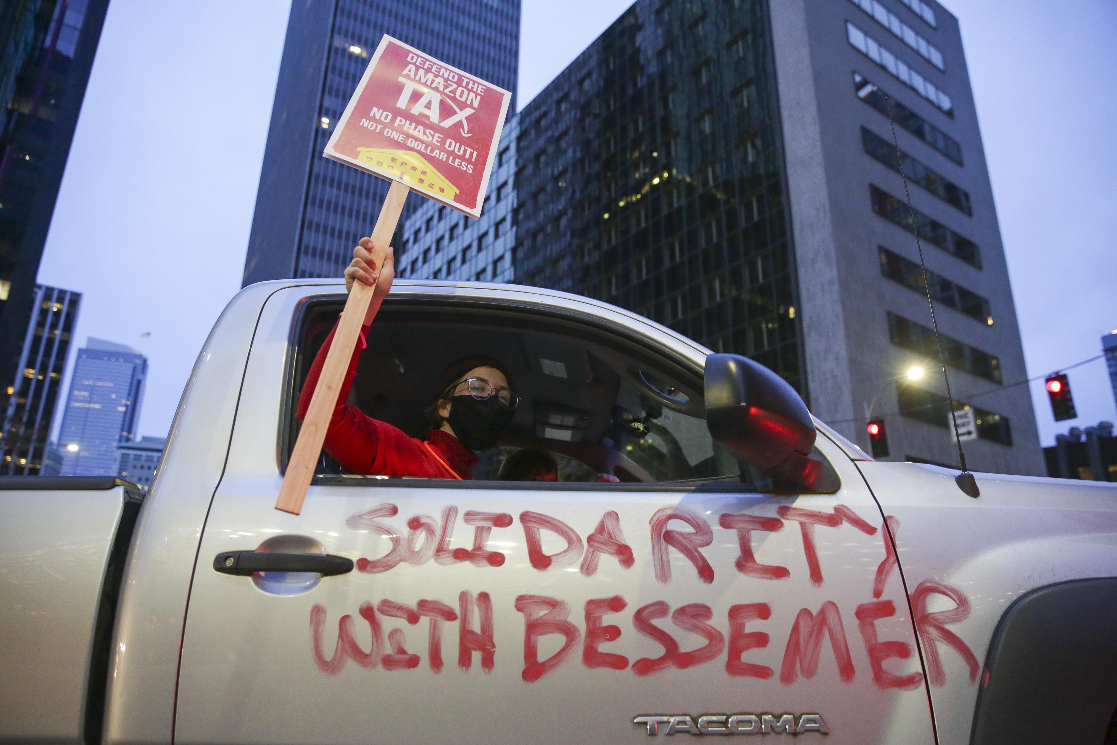 A demonstrator holds up a "Defend the Amazon Tax" sign through the passenger side window of a truck with "Solidarity With Bessemer" painted on the outside, part of a Tax Amazon car caravan.