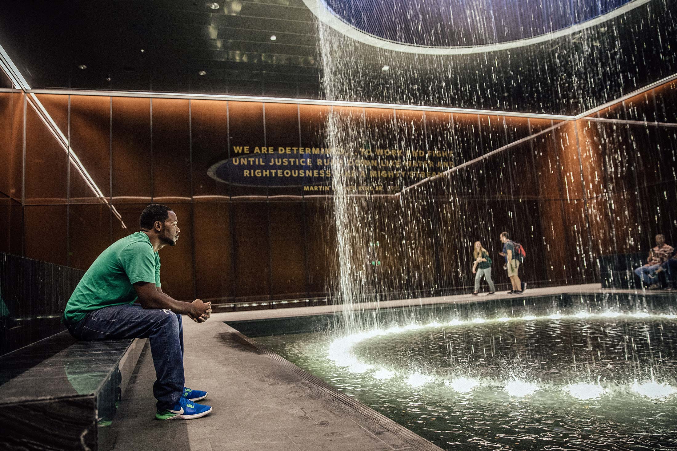 A black man sits on a bench and looks into a fountain in the National Museum of African American History and Culture.