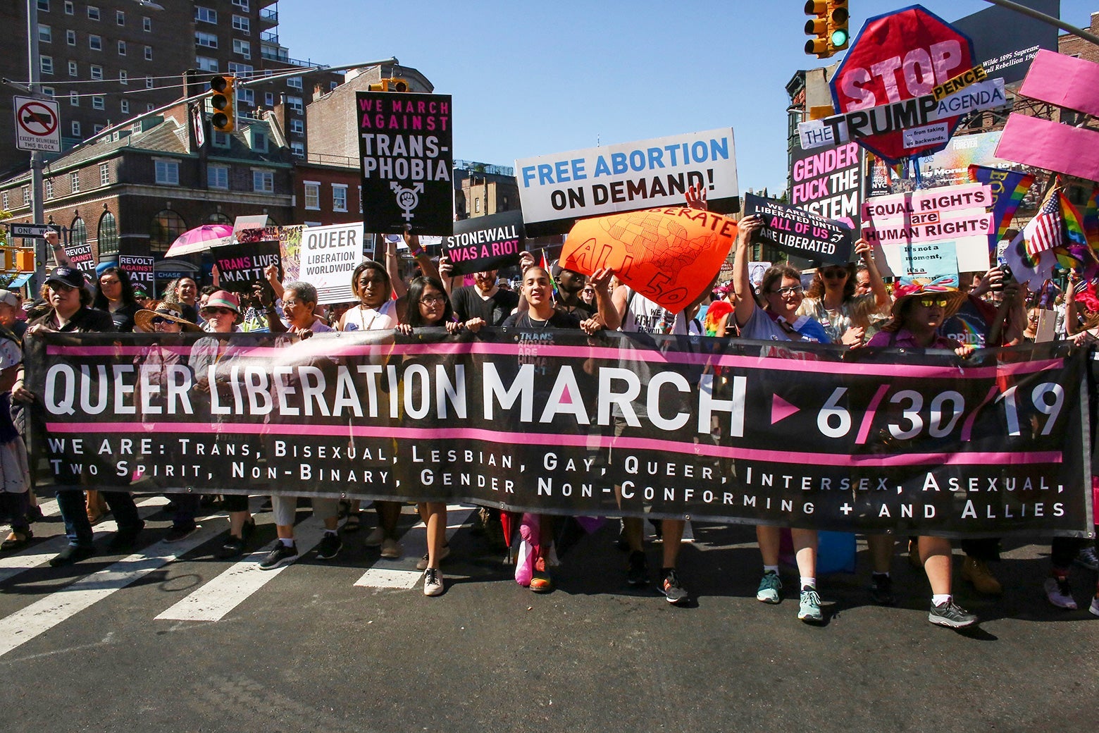 A crowd of protesters on a street carry signs and display a banner reading "Queer Liberation March."