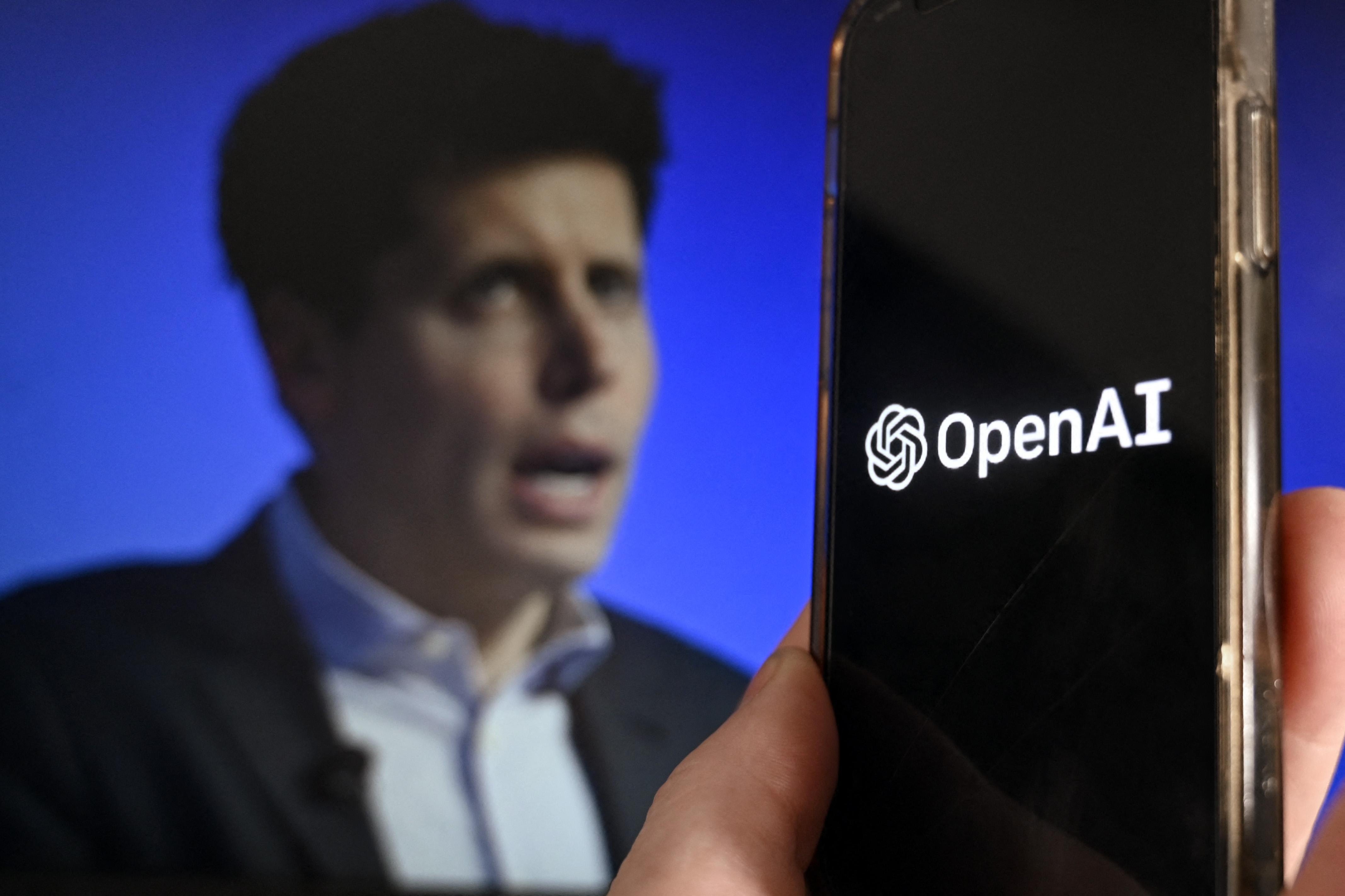 A smartphone displaying the OpenAI logo is held up in front of a background display of Sam Altman's face.