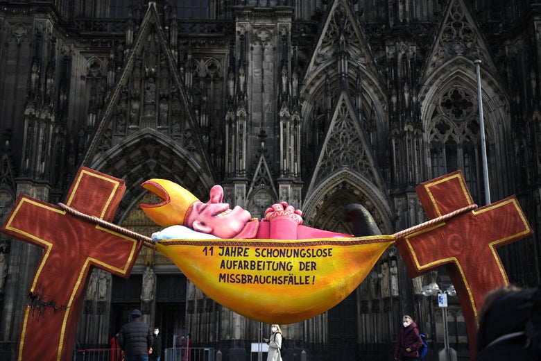 A sculpture depicts a cartoon bishop sleeping in a hammock strung between two crosses, which are cracking under the weight. The sculpture is in front of a Gothic cathedral.