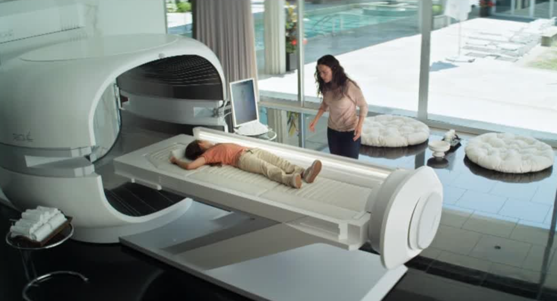 In a still from a movie, a woman looks at a small child about to go inside a medbed-looking device.
