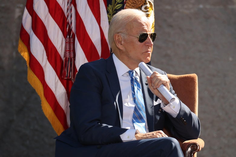Biden sitting in an armchair with his legs crossed and a rolled up piece of paper clutched in his left hand at the 10th anniversary celebration of the Martin Luther King Jr. Memorial. He is wearing aviator sunglasses and looking to his left with a stern expression. An American flag hangs from a pole behind him.