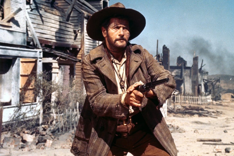 Eli Wallach dressed in a cowboy hat holds a gun out pointed in front of him as he stands in a town of ruins that's still smoking. 