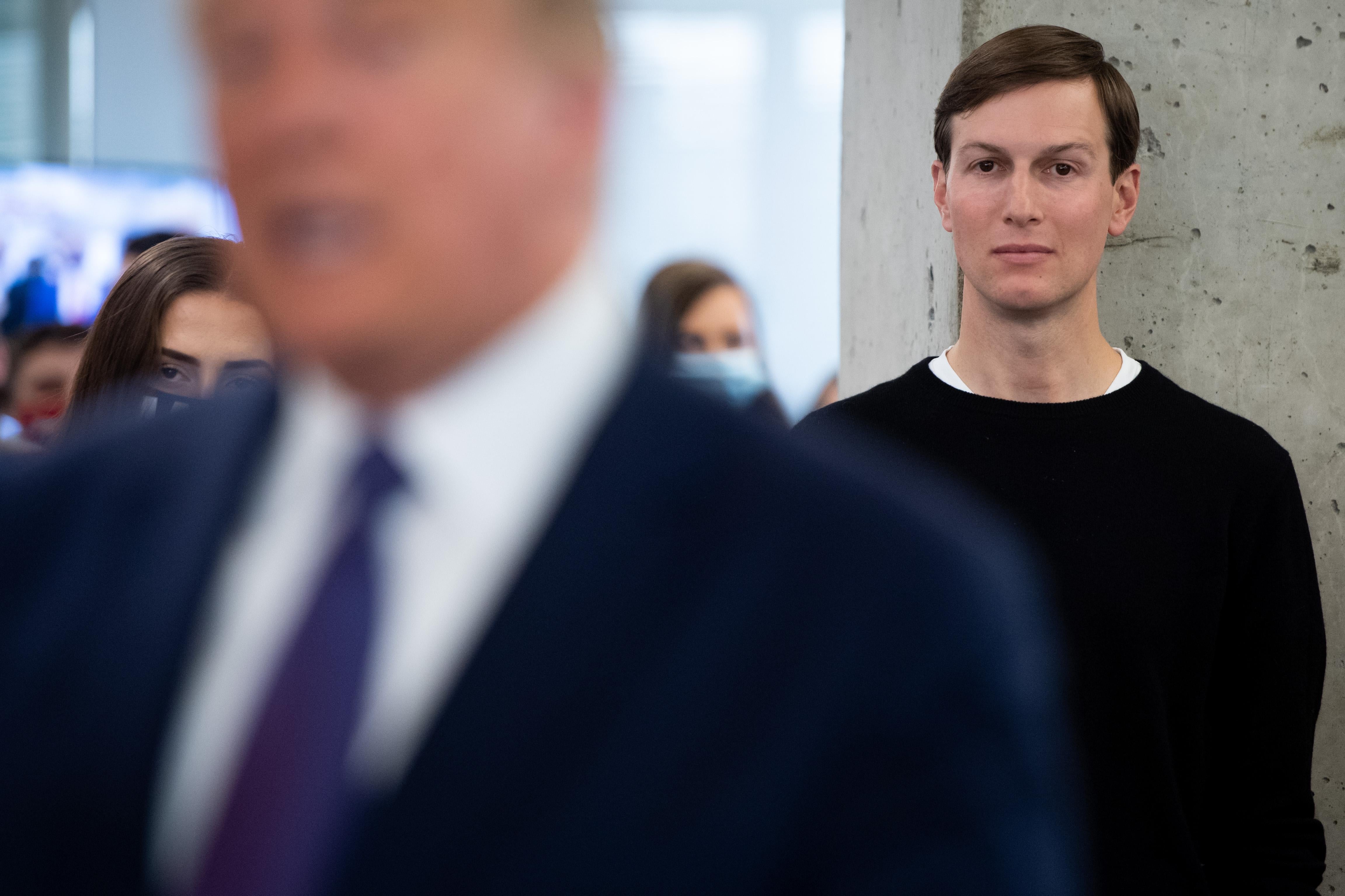 A blurry figure of Donald Trump fills the left foreground while Jared Kusner, in a dark crewneck, gazes blankly while standing against a concrete wall in the right rear. 