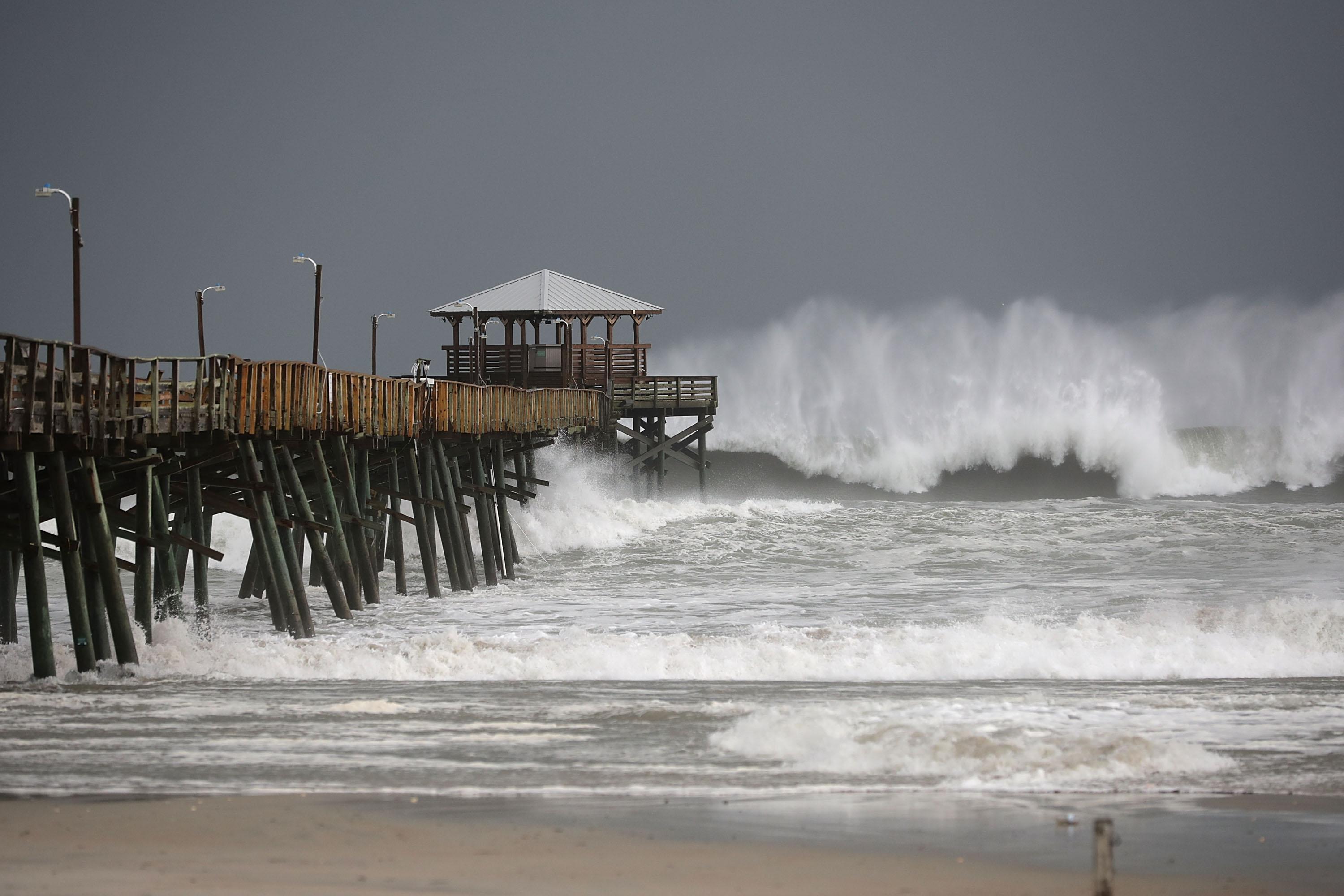 High waves crash around a pier in front of a gray, stormy sky.