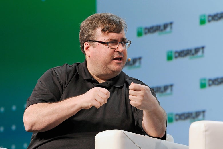 A man sitting on a stage raises his hands as he speaks.