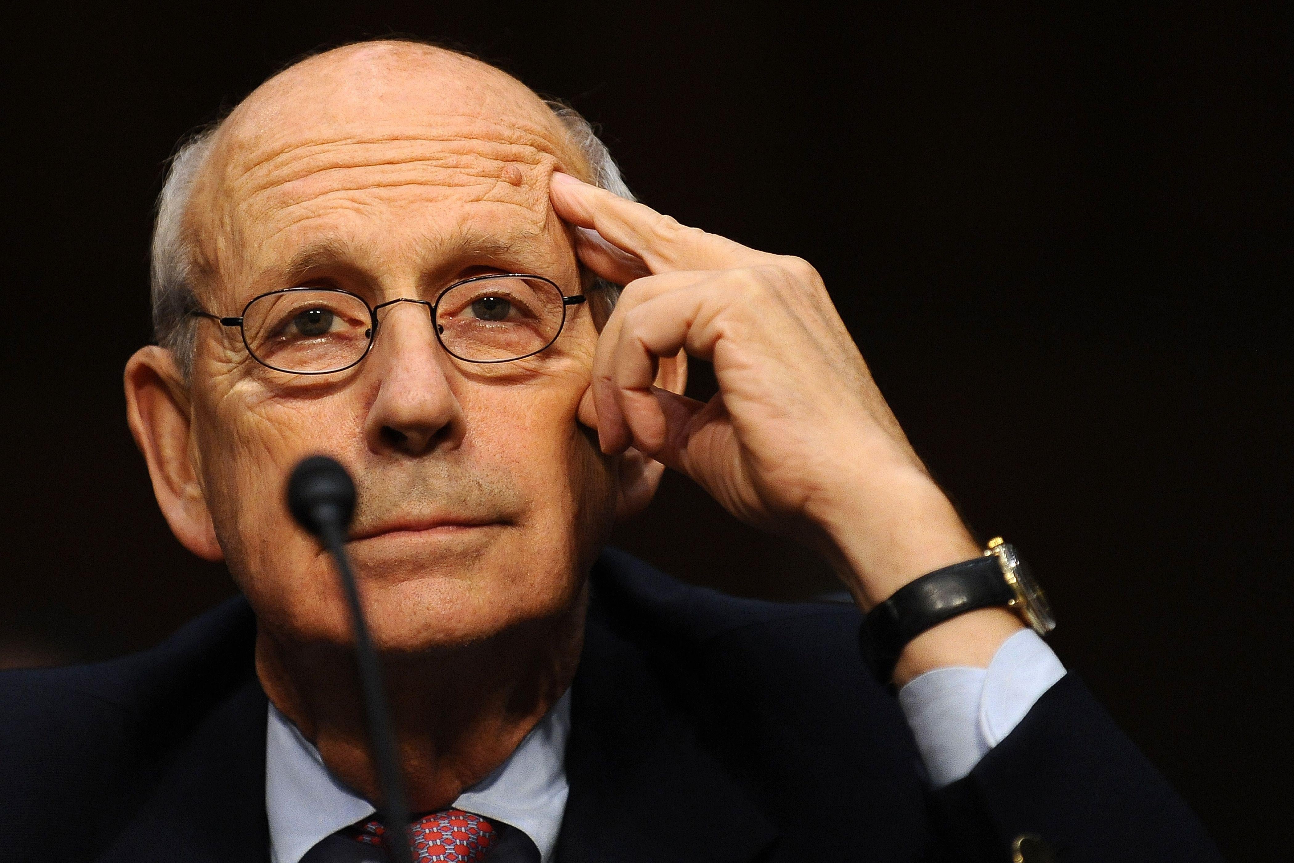 Breyer touches his temple as he smiles mildly sitting in front of a mic