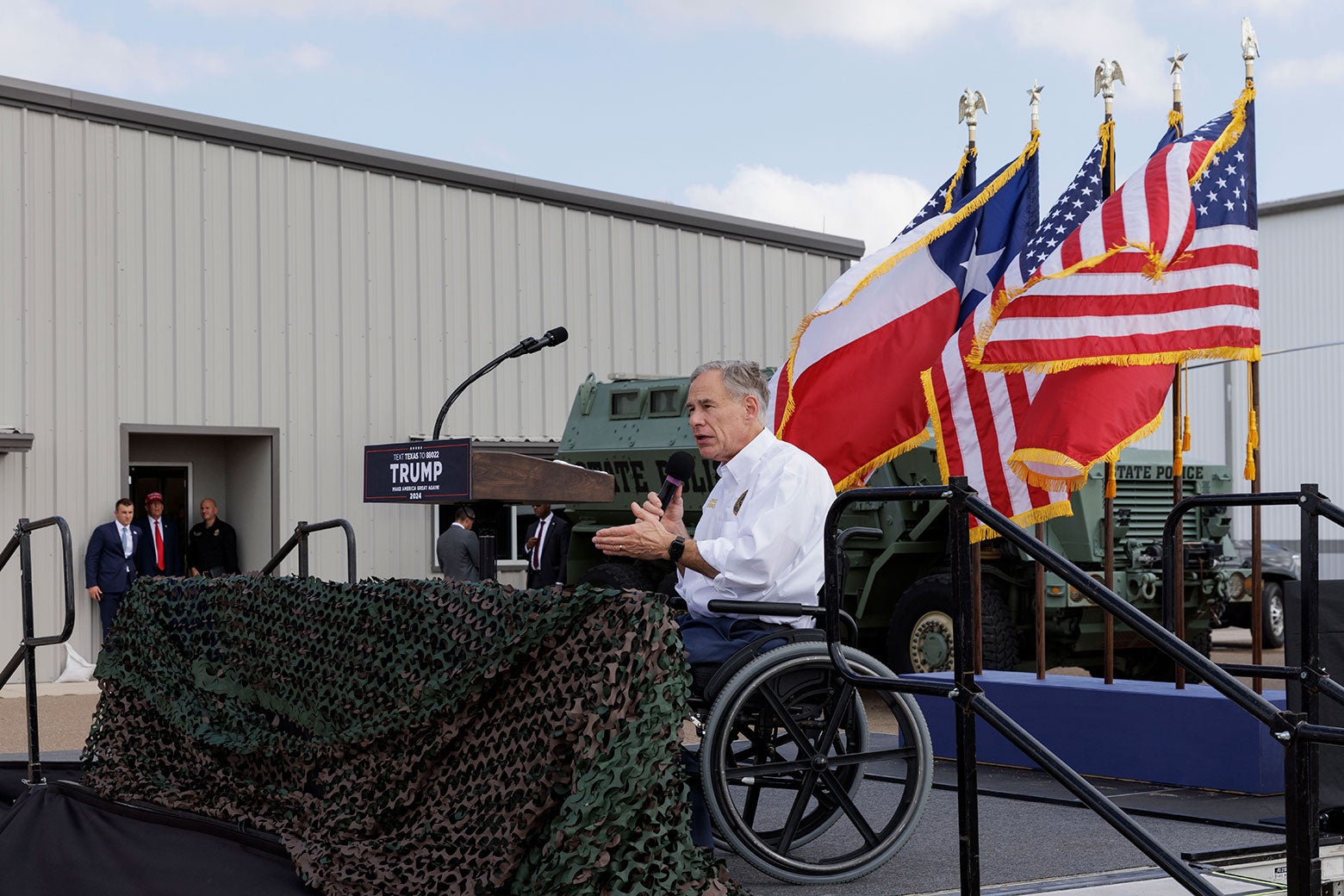 Abbott, in his wheelchair, talks to a crowd from a stage with Texas and American flags behind him.